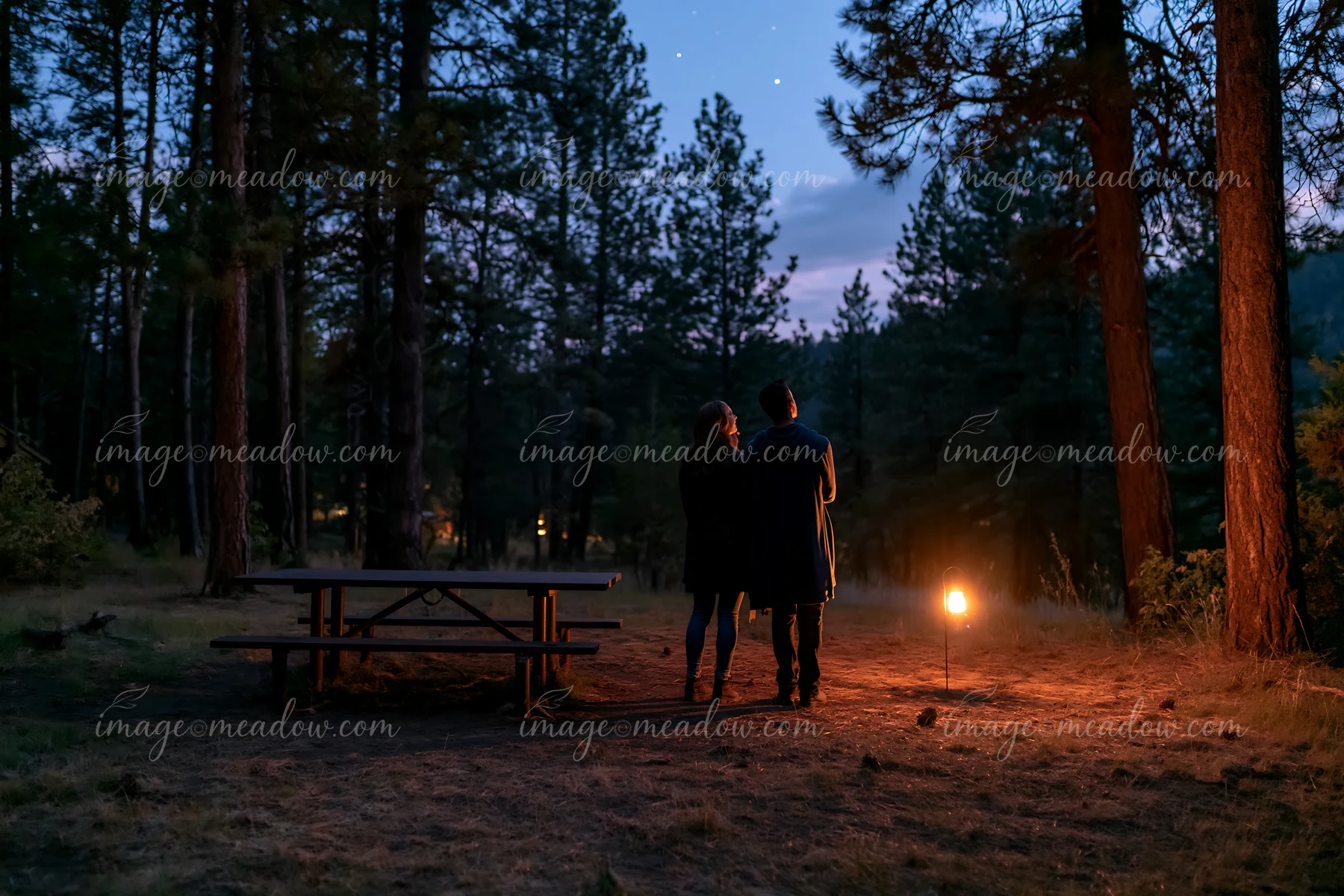 A couple stargazing in a national park stands together near a lantern as night falls.