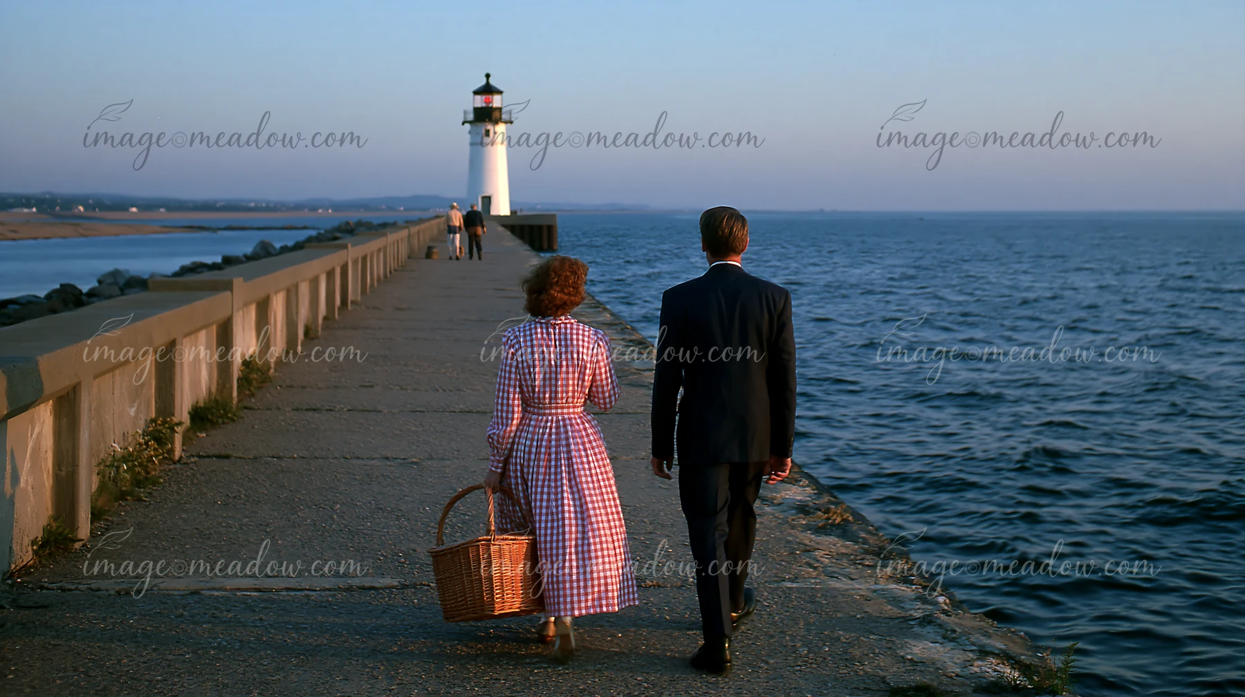 A couple walks along a coastal breakwater toward a white lighthouse beside the sea.