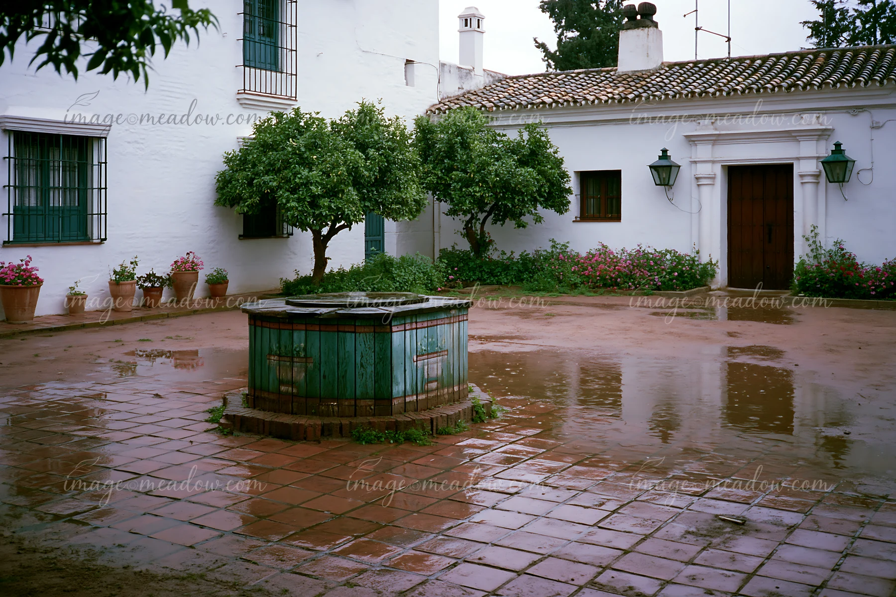 A courtyard features a stone well surrounded by wet tiles after rain.