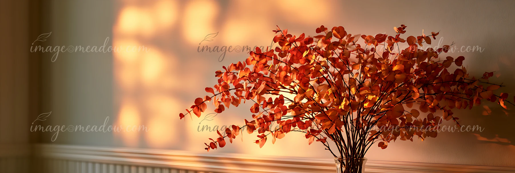 A decorative branches arrangement with orange leaves sits on a table while golden light from a window creates soft shadows on the wall in a living room setting