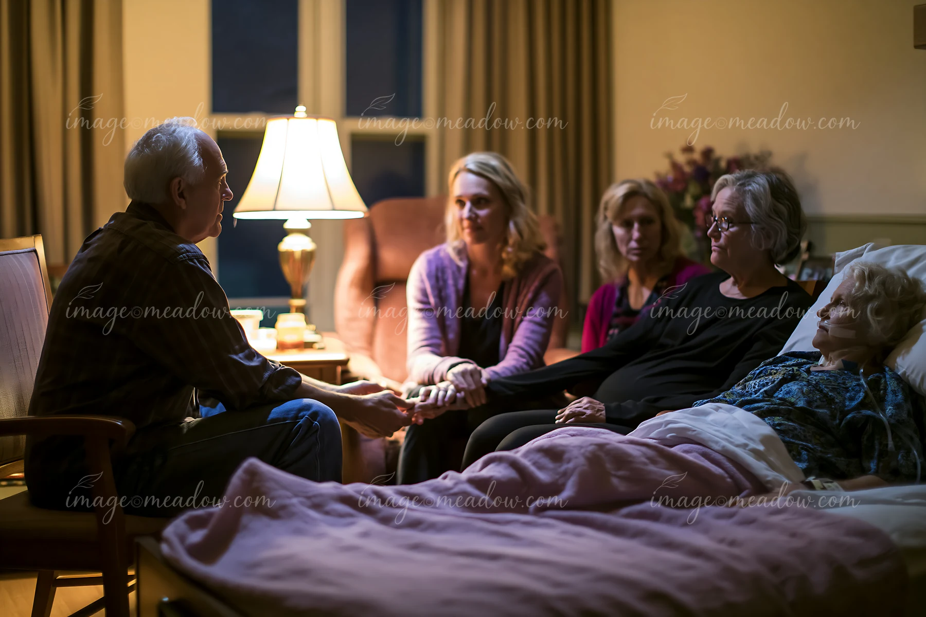 A family sits closely together, holding hands while sitting beside a hospice patient.
