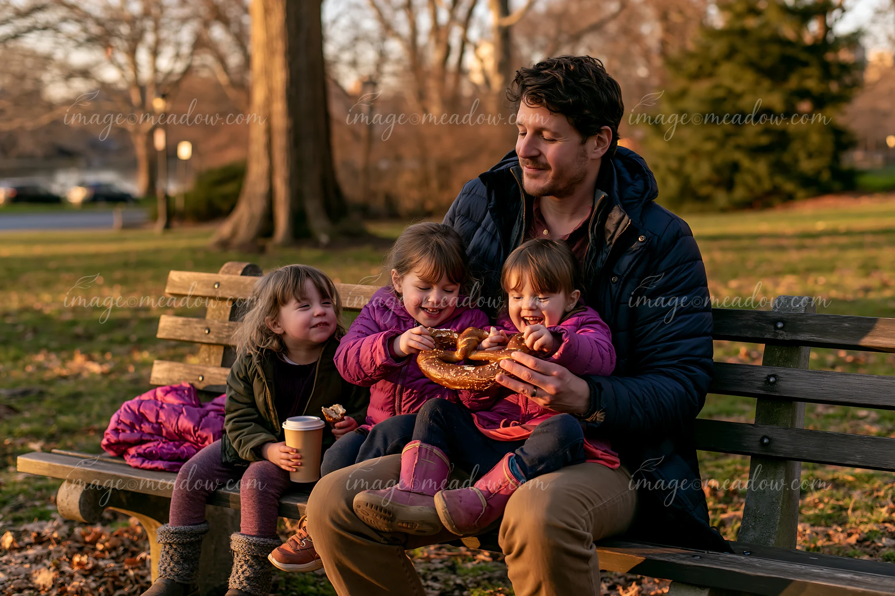 A father and children sharing a giant pretzel while sitting on a park bench during sunset.