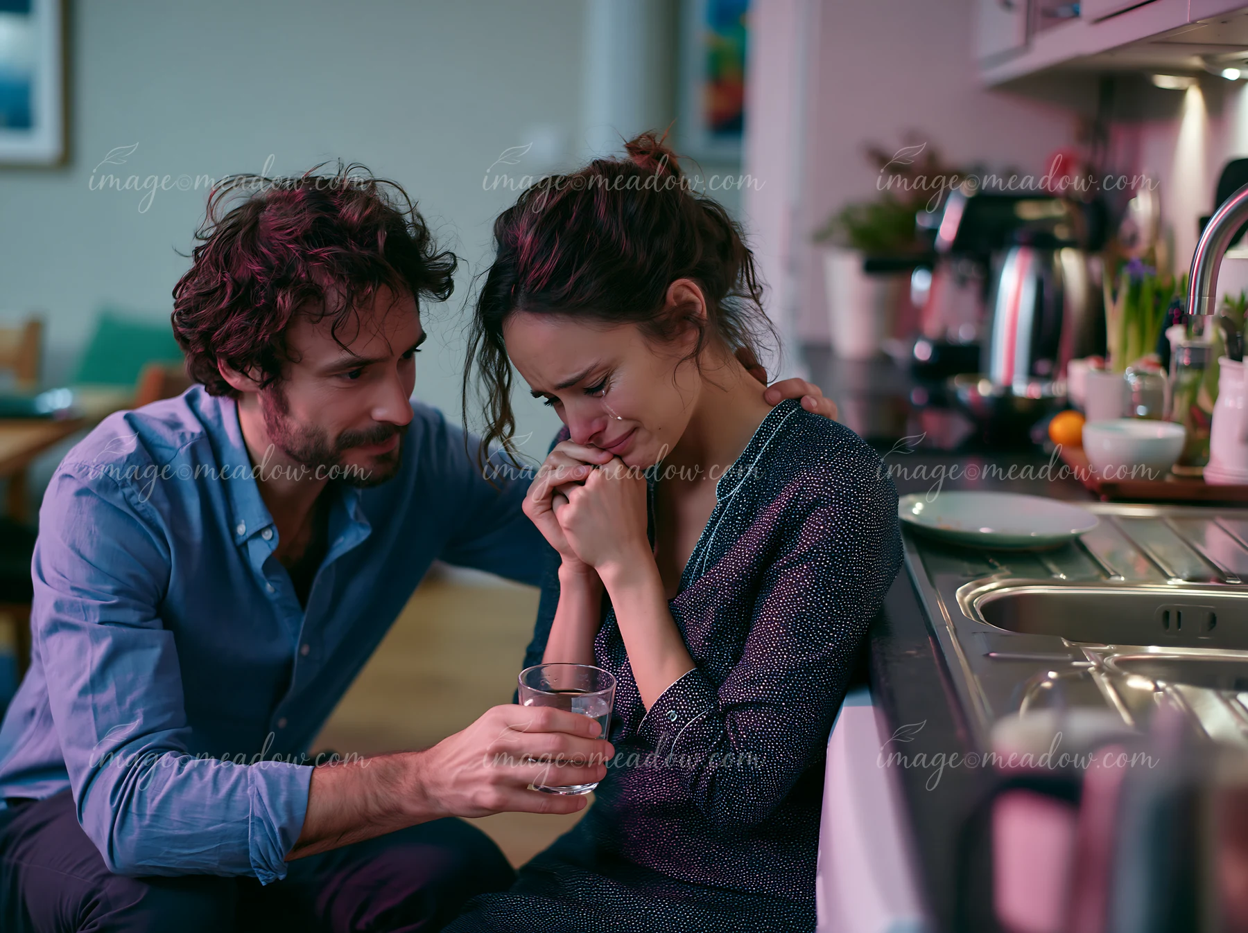 A friend comforts a crying woman in a home kitchen while offering her a glass of water.