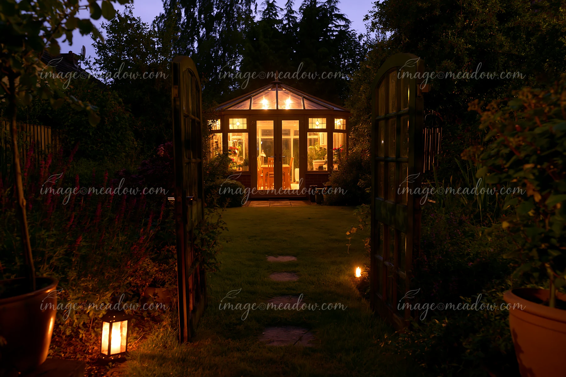 A garden conservatory stands lit from within, surrounded by plants and trees.