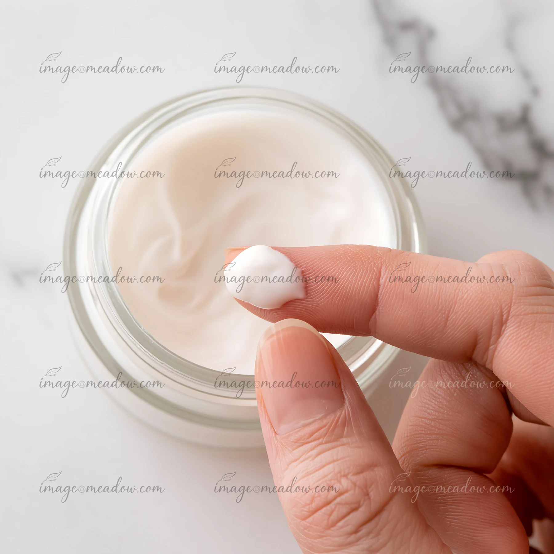 A hand applies a small amount of cream from a jar to the fingertip.