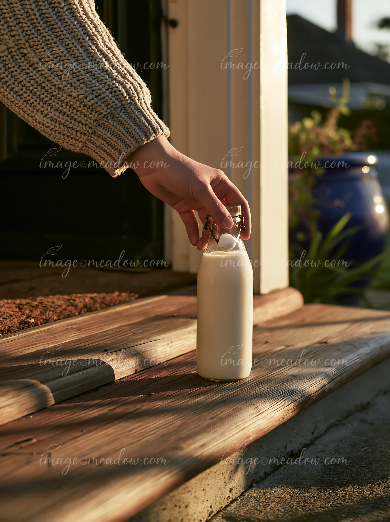 A hand reaches for a glass milk bottle on a doorstep at sunrise.