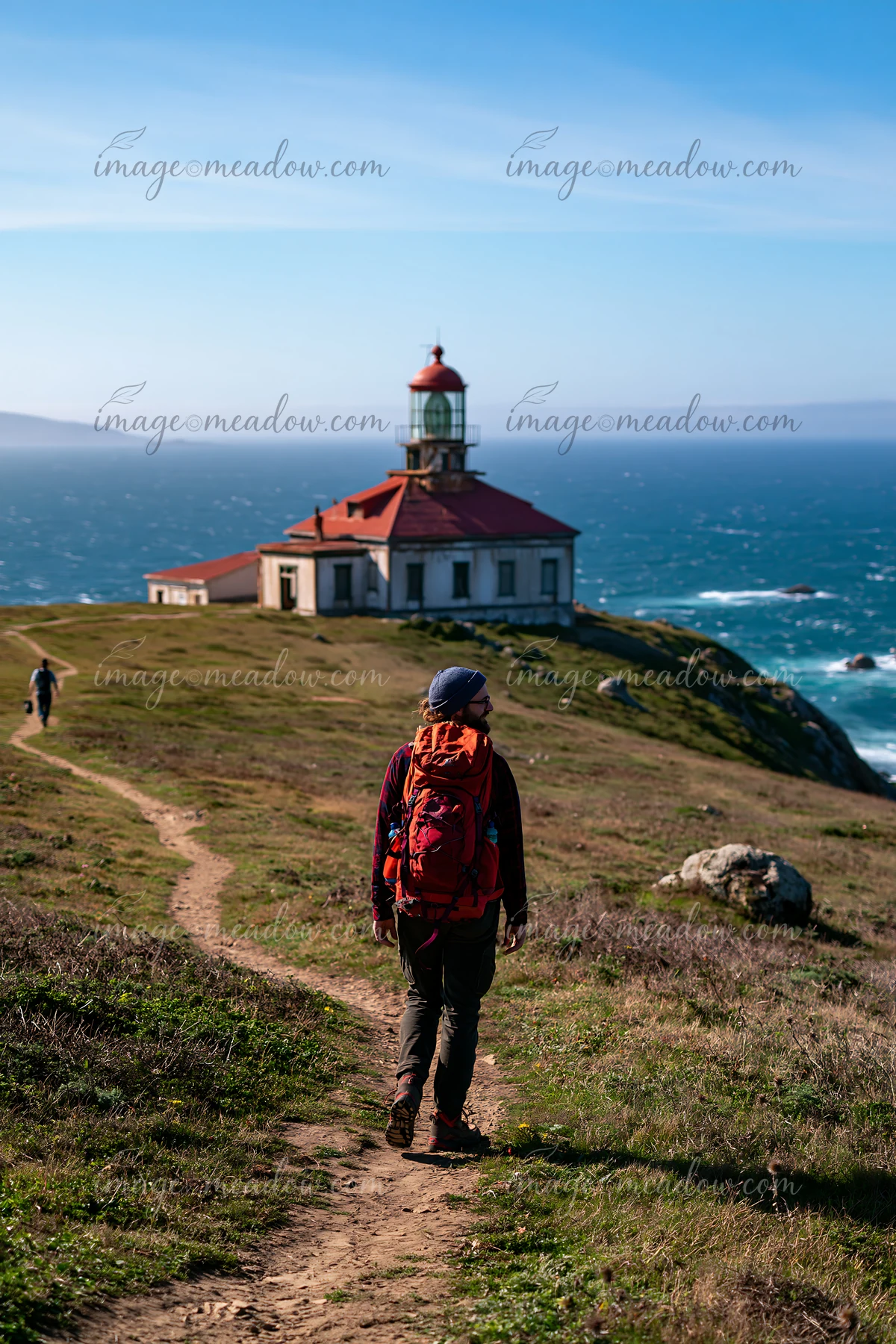 A hiker walks along a dirt path toward an abandoned lighthouse on a remote coast.