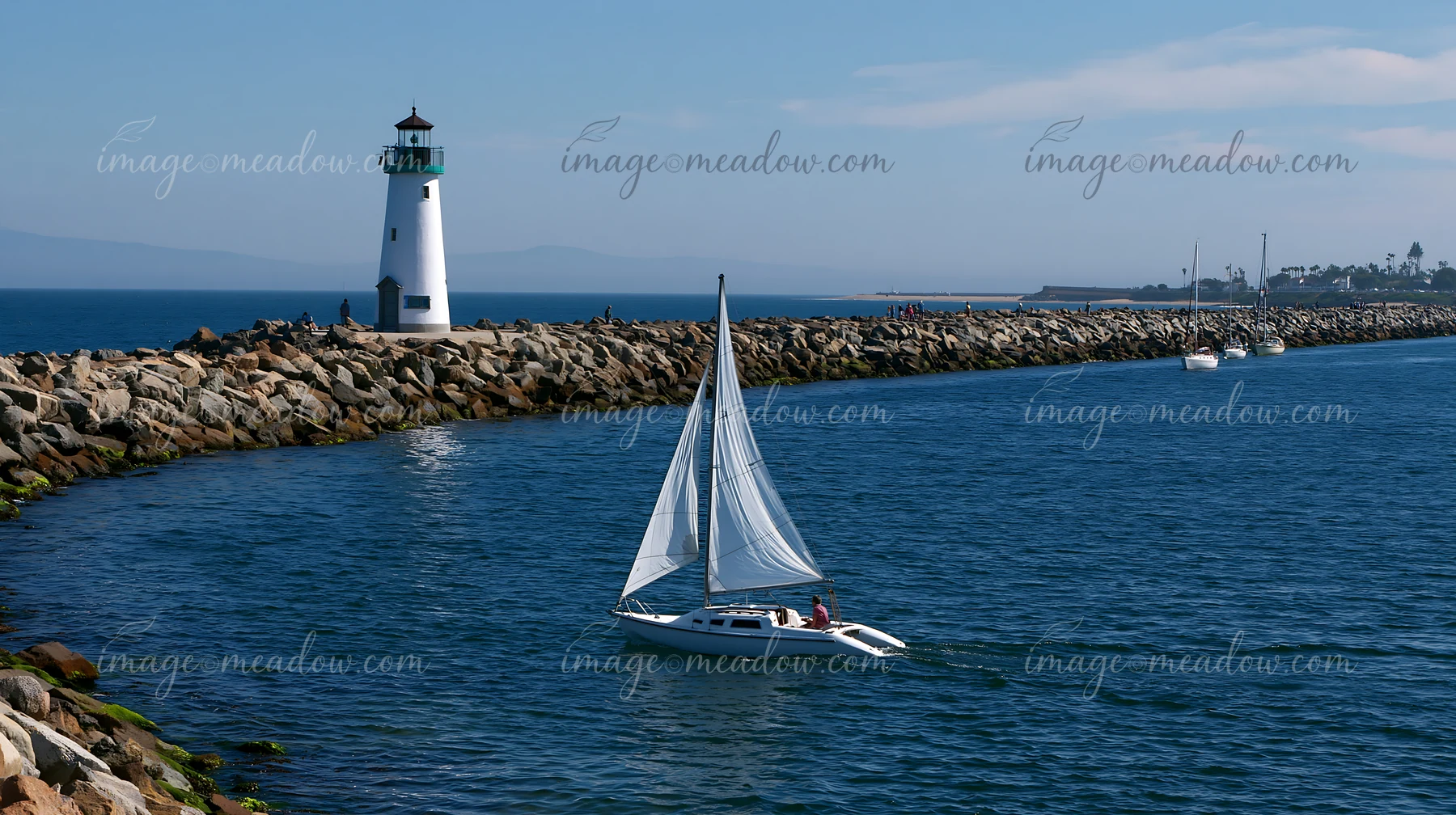 A lighthouse stands tall at the end of a rocky pier, while sailboats glide through the calm waters.