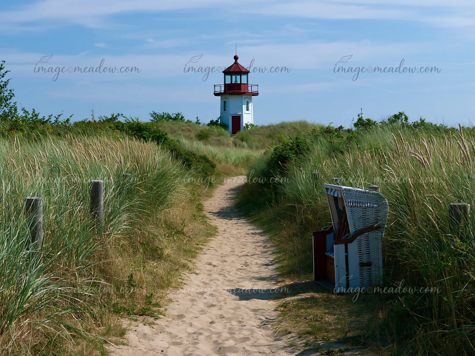 A lighthouse stands tall at the end of a sandy path, surrounded by coastal dunes and tall grass.