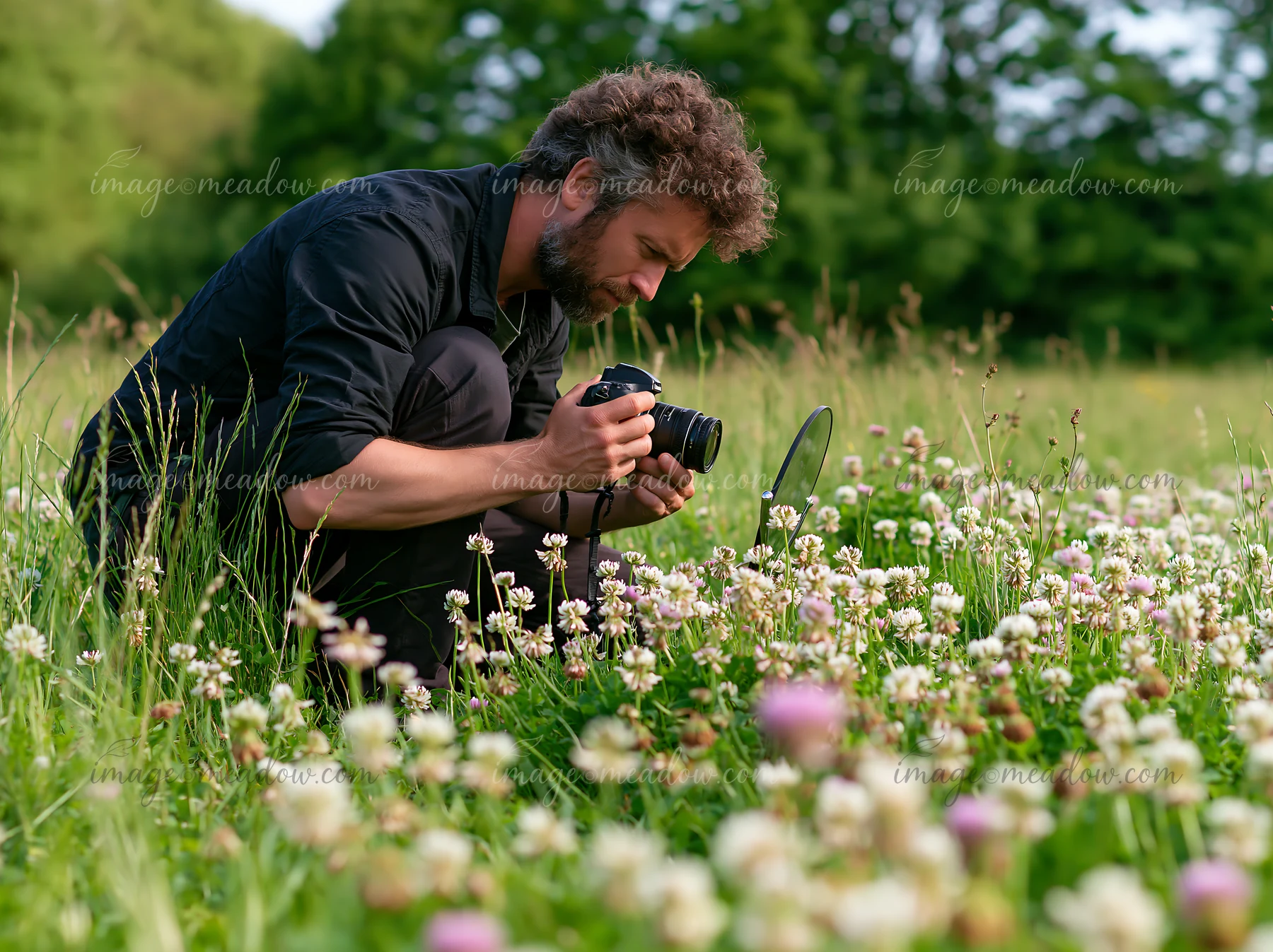 A man kneels in a green field filled with wildflowers, capturing images with his camera.