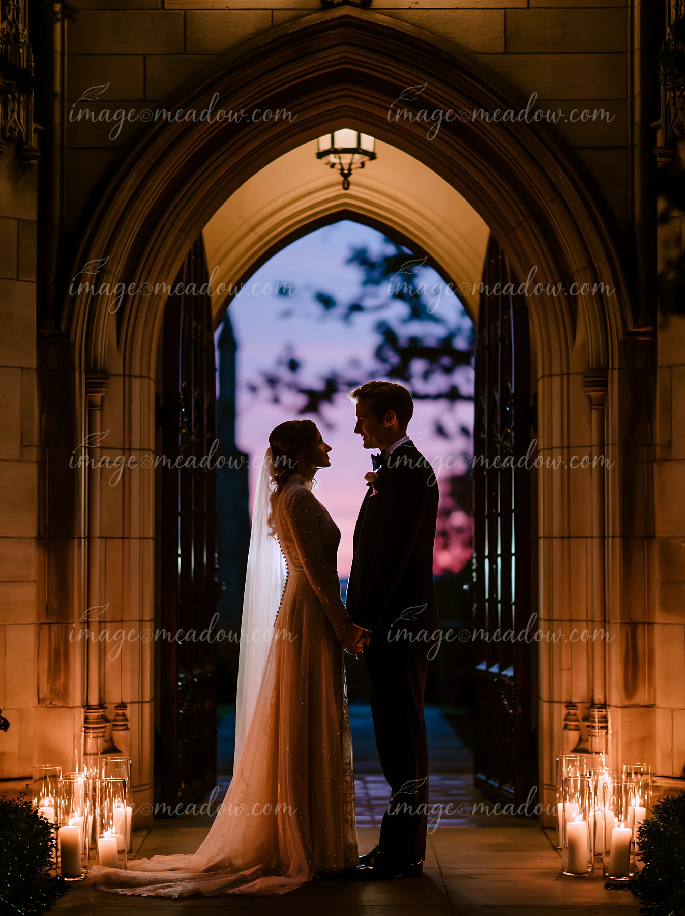 A newlywed couple stands under an arch at a church doorway while holding hands.