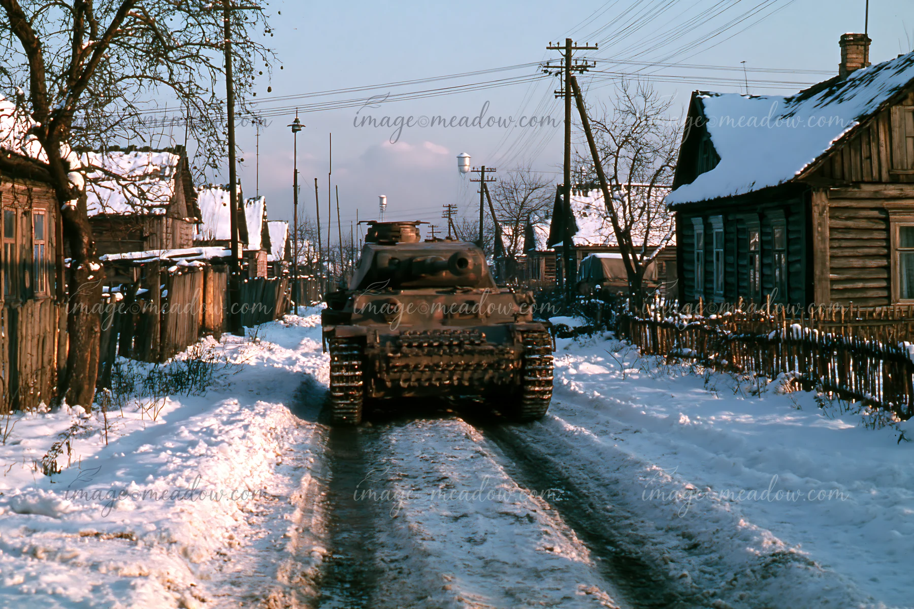 A panzer tank in winter snow drives down a narrow road lined with wooden houses.