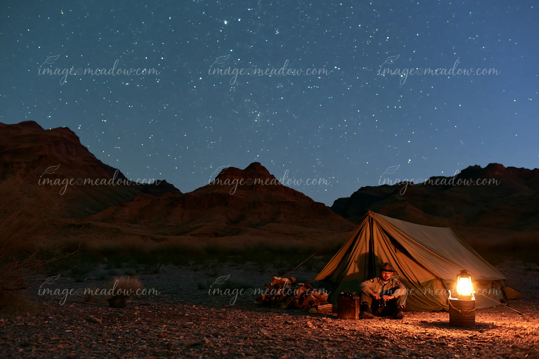 A person sits by a lit lantern at a desert campsite with a tent beneath a sky full of stars.