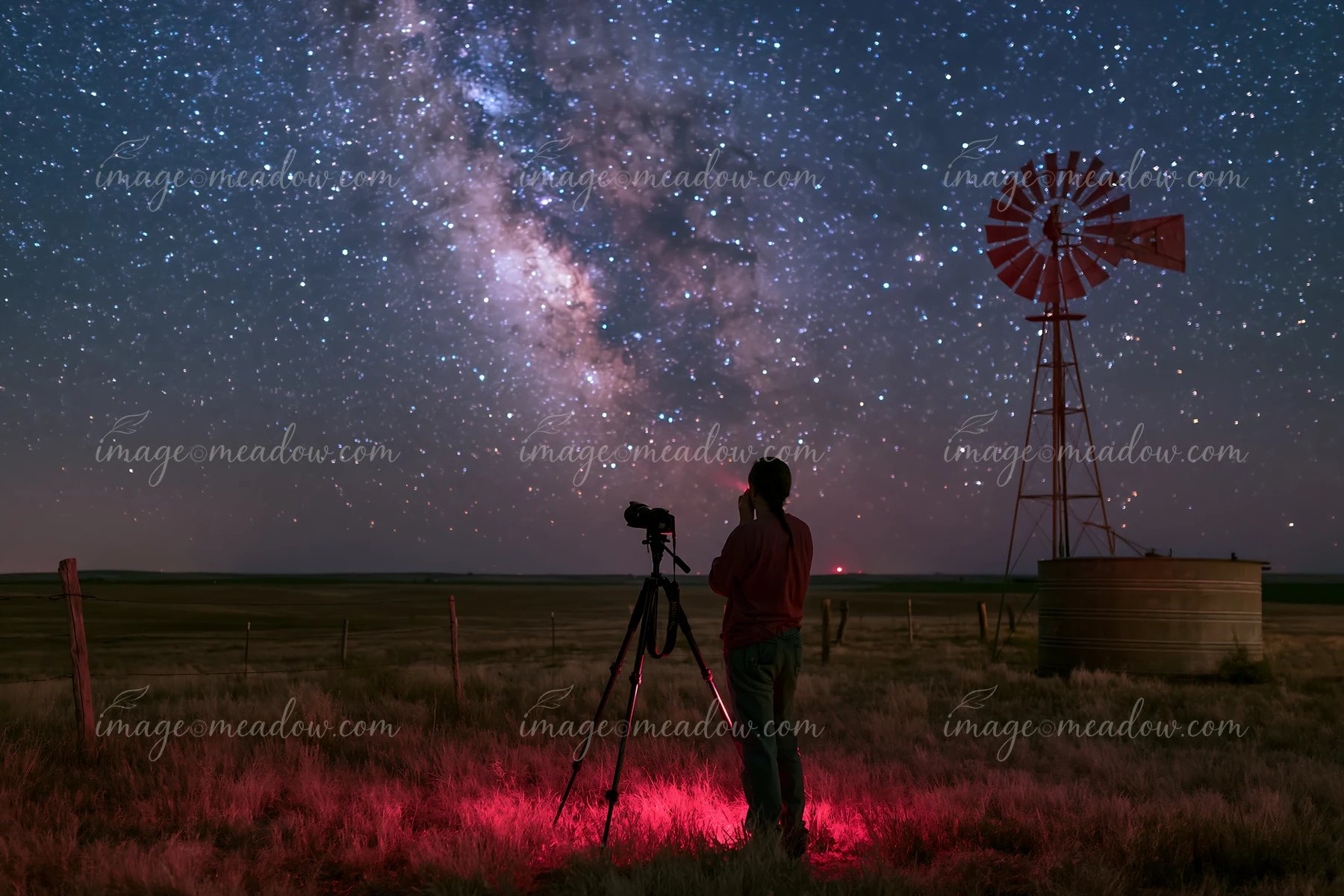 A person using a camera under the Milky Way stands next to a windmill in a rural field at night.