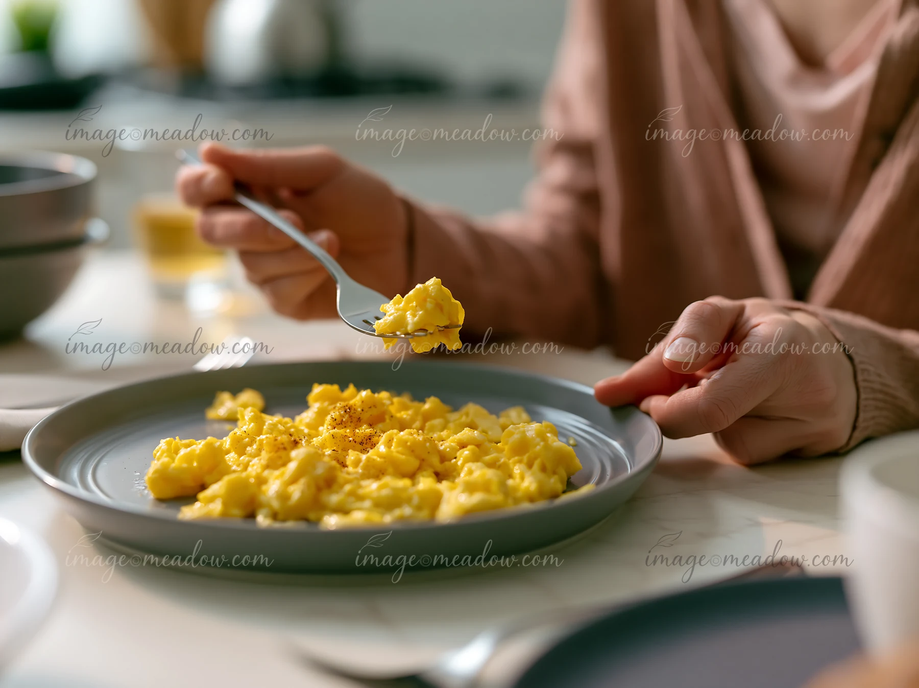 A person with limited hand mobility using utensils to eat scrambled eggs from a plate in a kitchen setting.