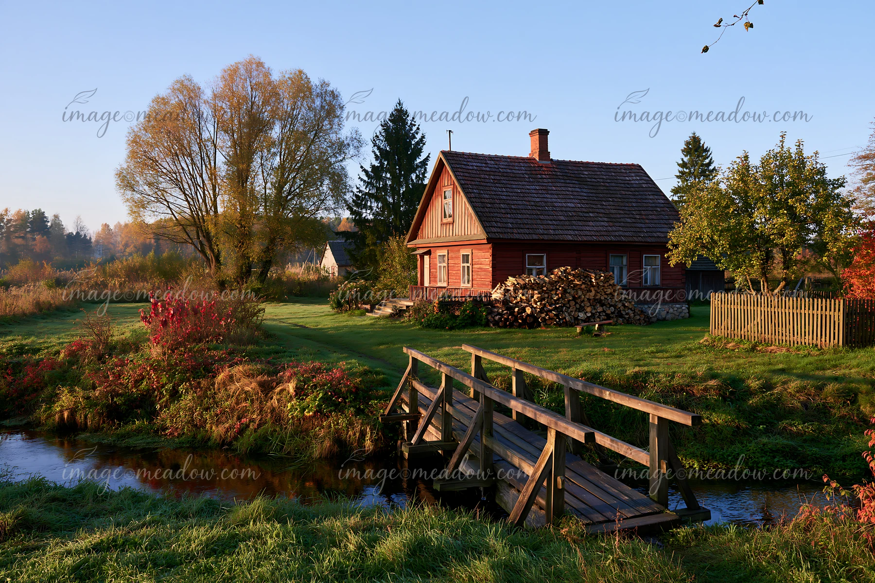 A rural cottage with firewood stack is surrounded by lush greenery and a small stream.