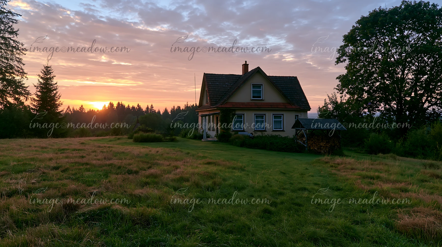 A rural cottage with firewood stack stands in a grassy field during sunset.