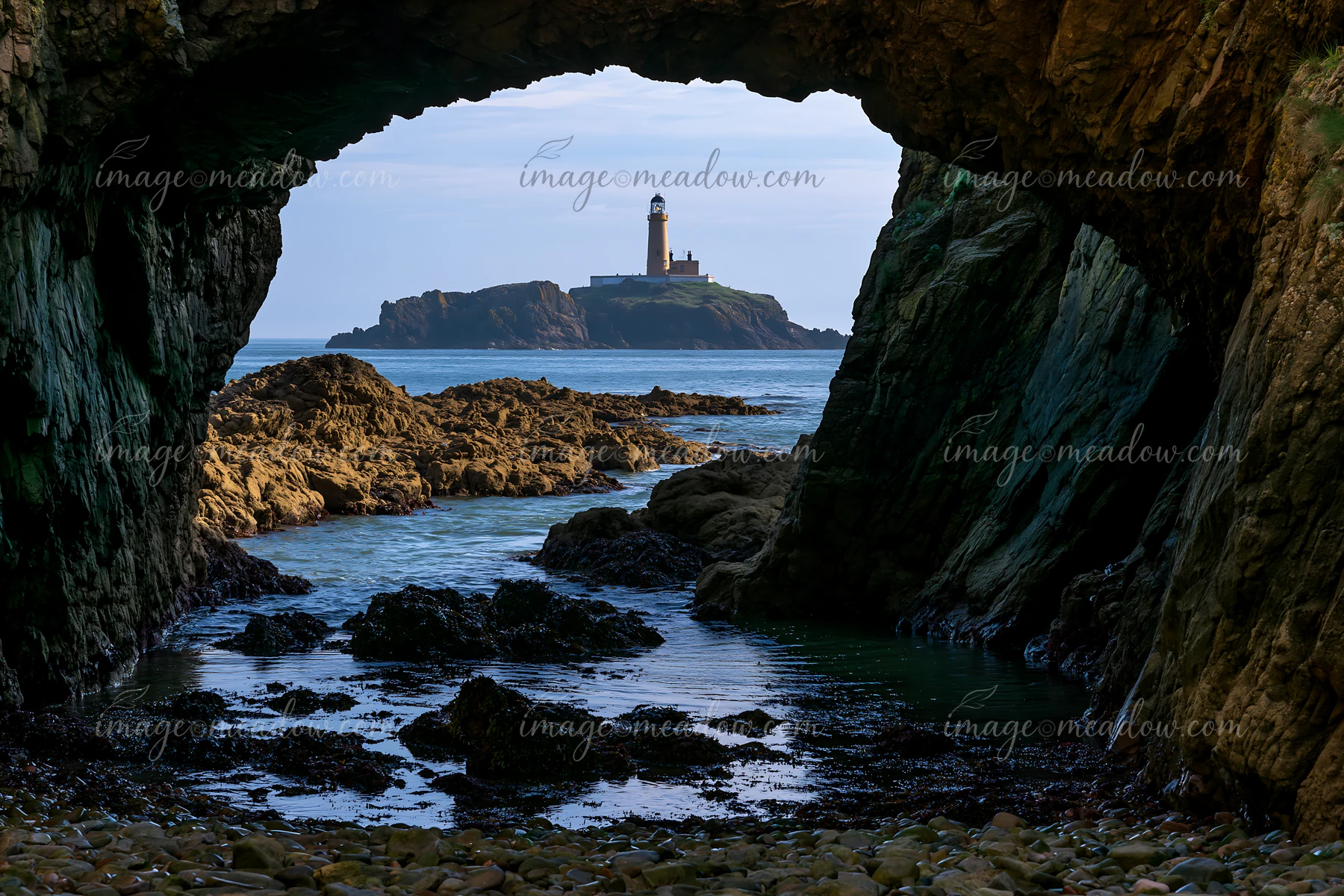 A sea cave frames a view of the coastline and a lighthouse situated on a distant island.
