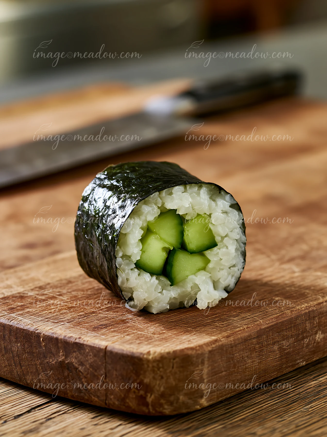 A single cucumber maki roll sits on a wooden cutting board.