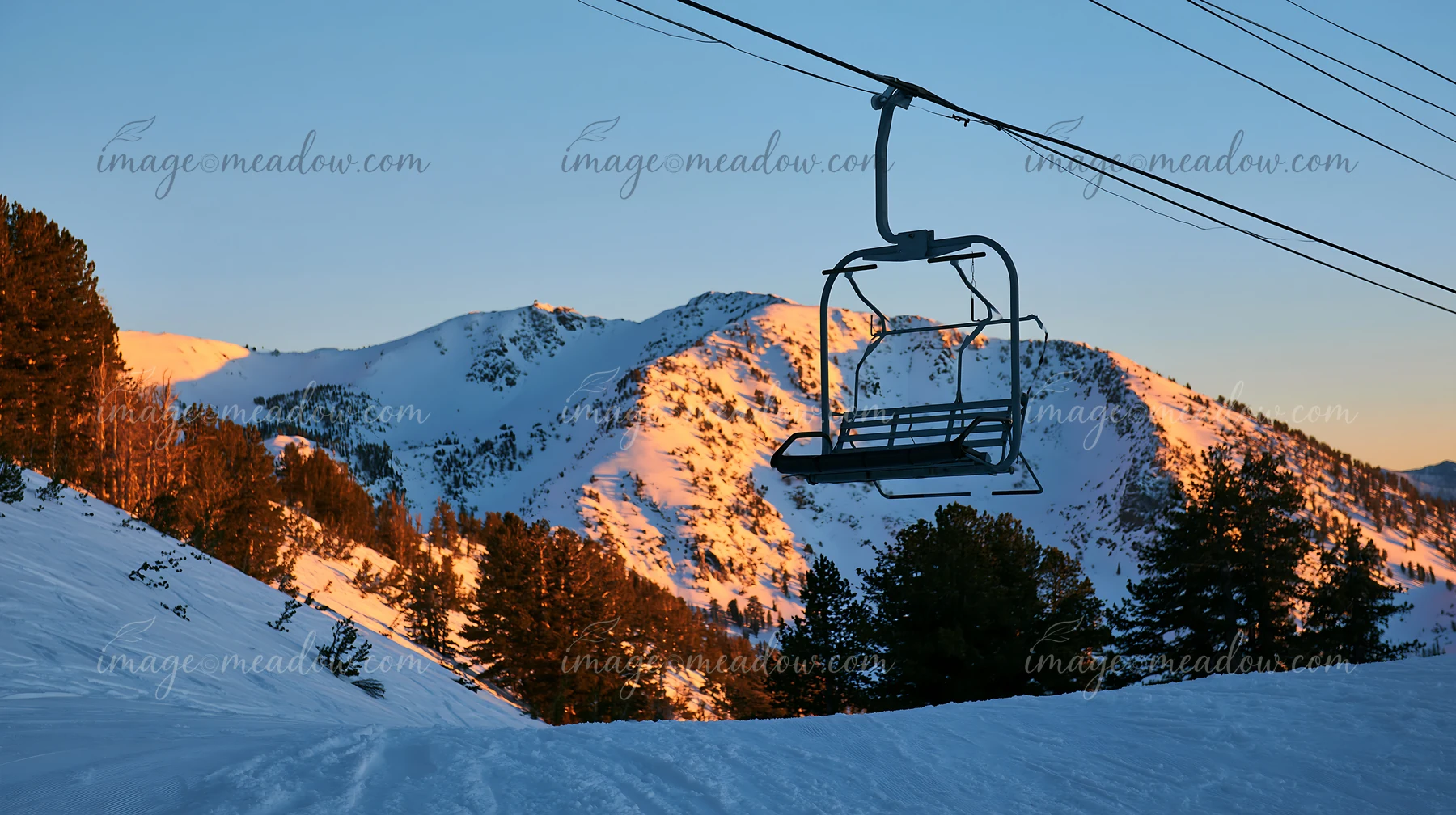 A ski lift stands still in a snowy landscape as the sun sets, casting golden light on the surrounding mountains.