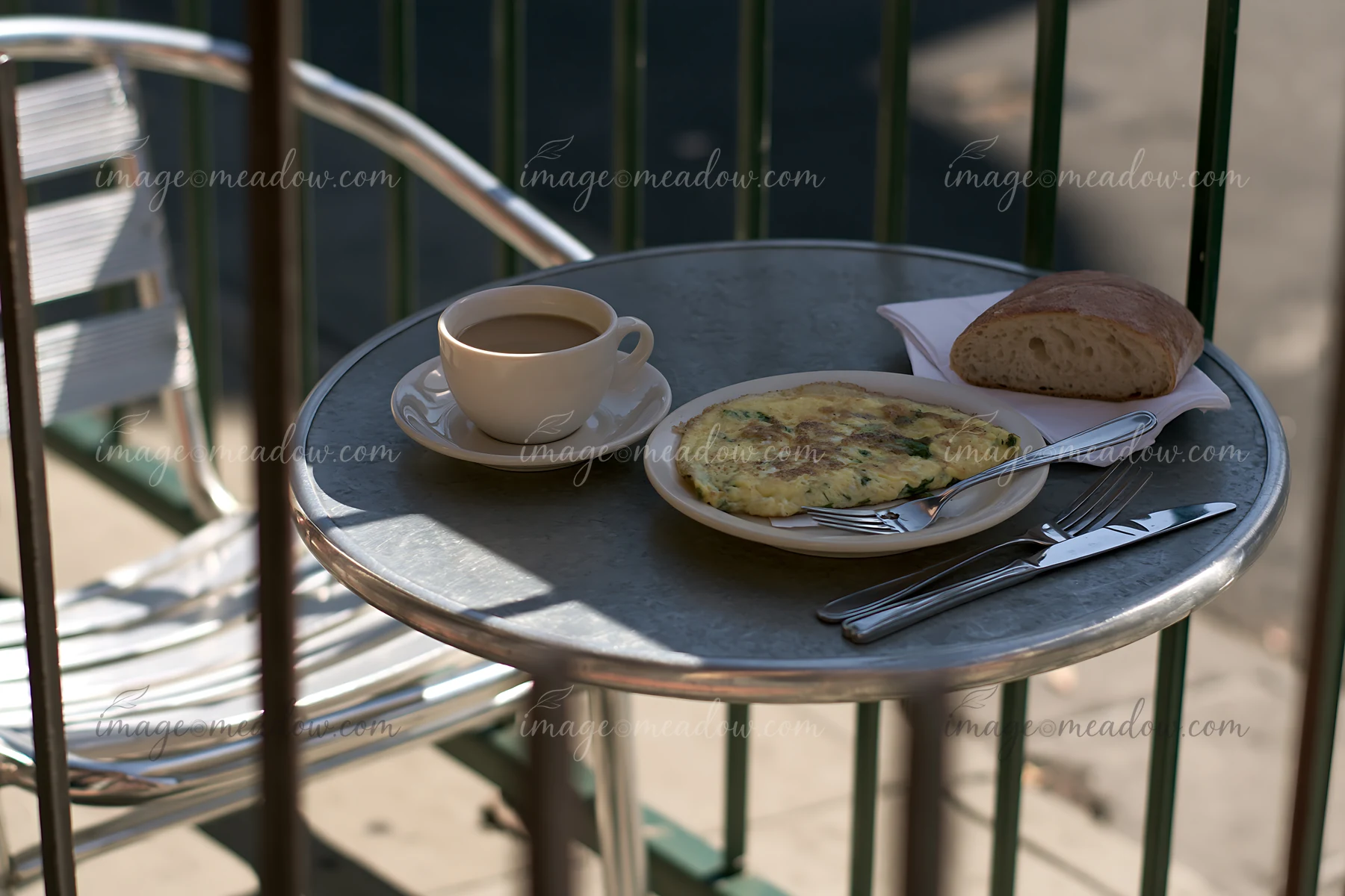 A small cafe table holds a cup of milk coffee, a plate with an omelet, and a piece of bread.