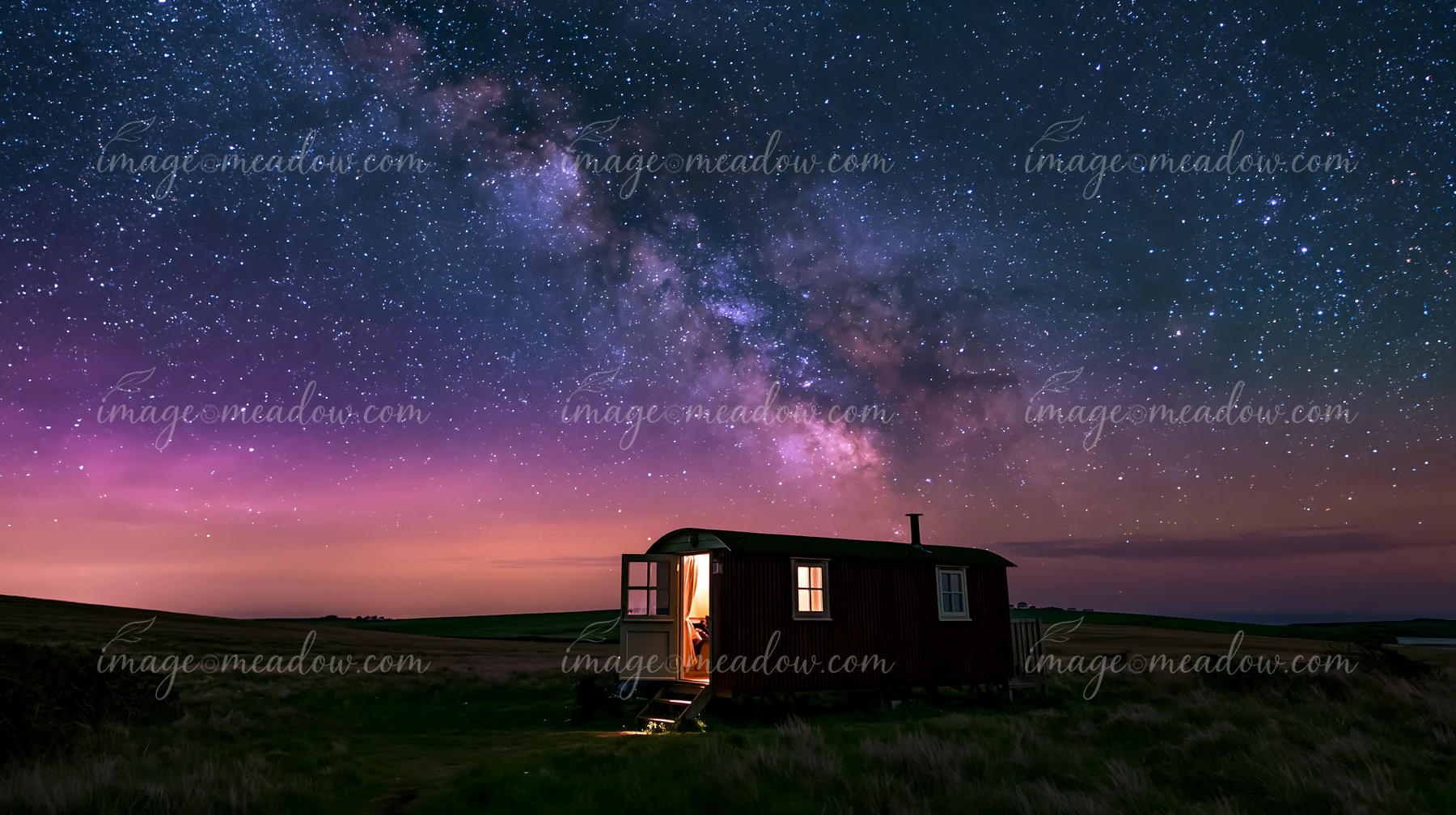 A small home sits in a vast wilderness with a bright light glowing from the window.