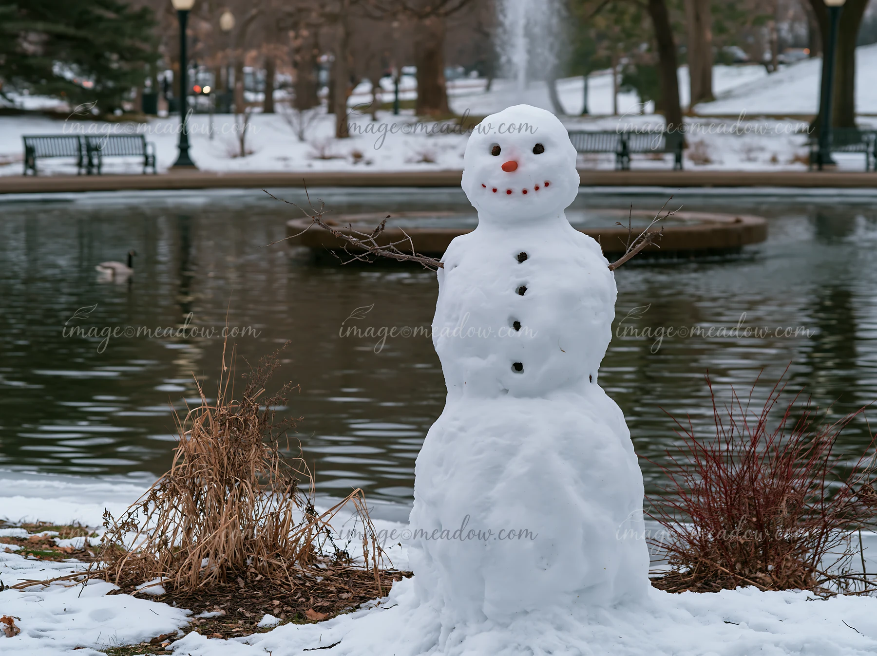 A snowman stands in a city park next to a frozen pond.