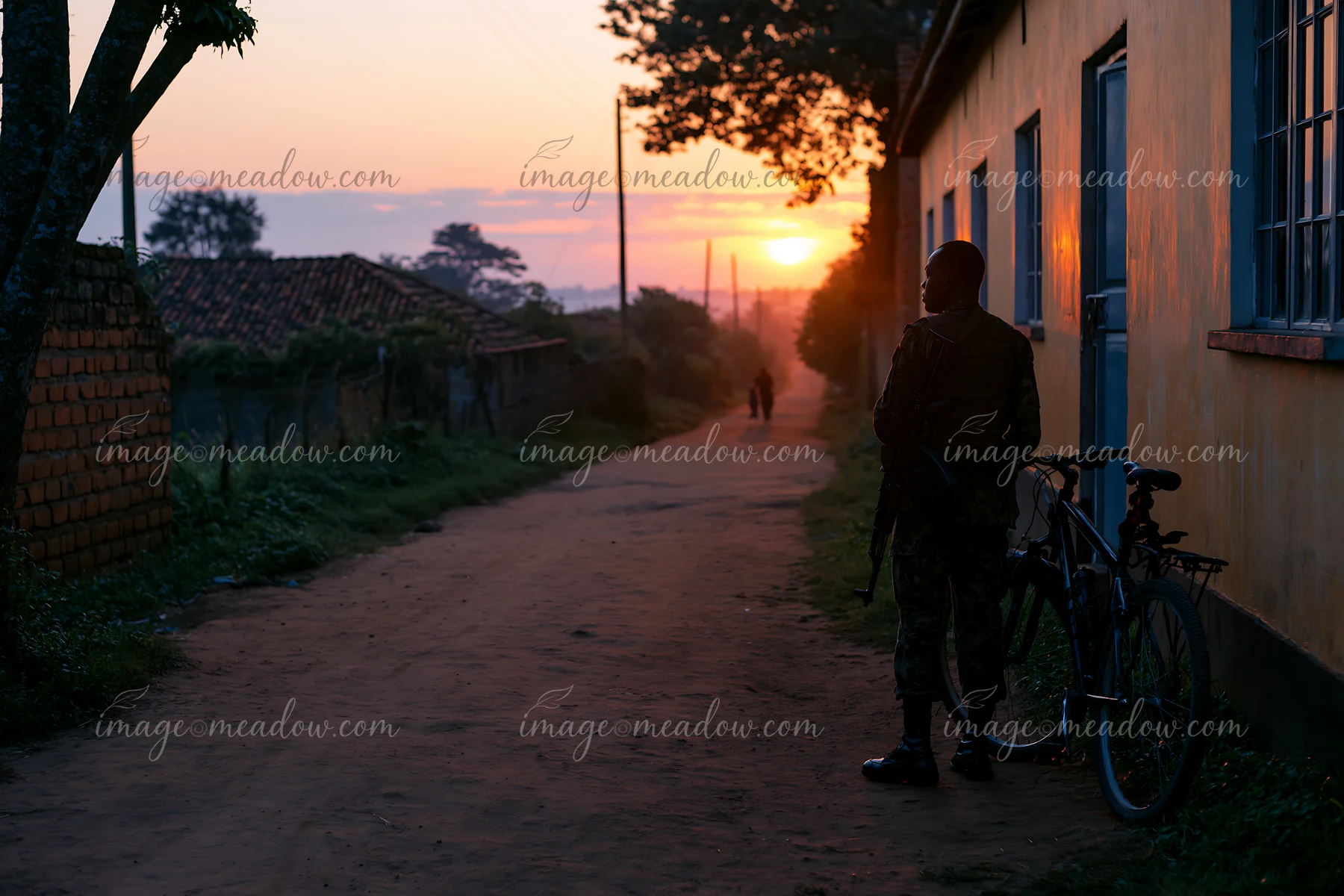 A soldier stands by a bicycle on a dirt road as the sun rises, with trees lining the path and a light fog in the air.