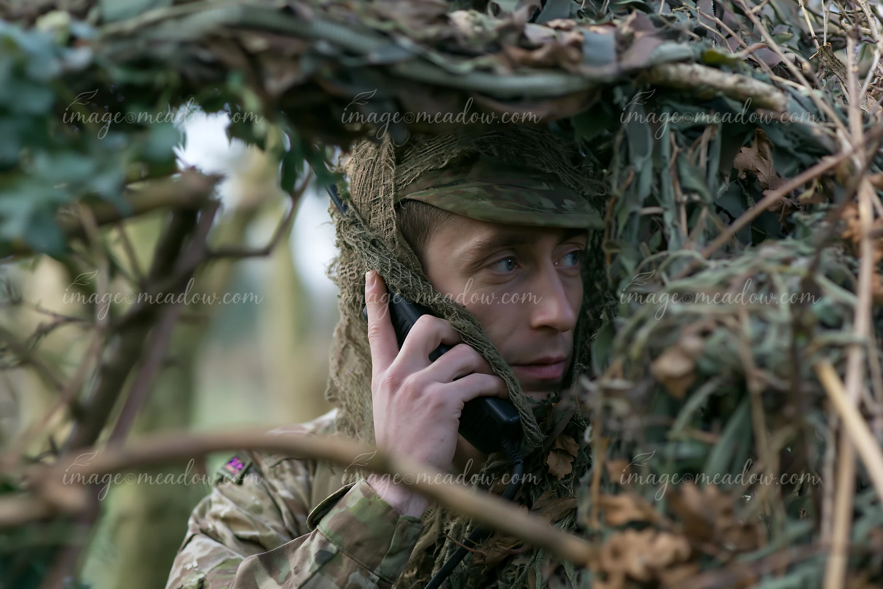 A soldier under camouflage netting communicates on a mobile phone while hidden in a natural environment.