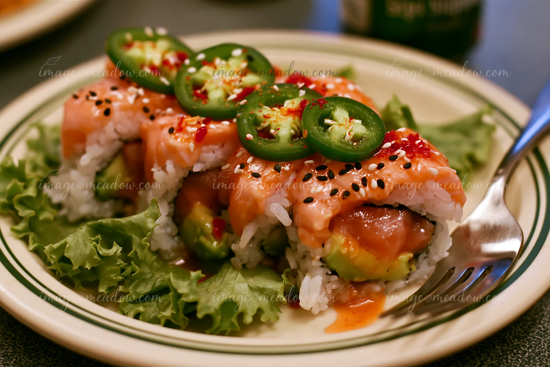 A spicy salmon sushi roll with chili placed on a bed of green lettuce, garnished with sesame seeds and sliced jalapeños.