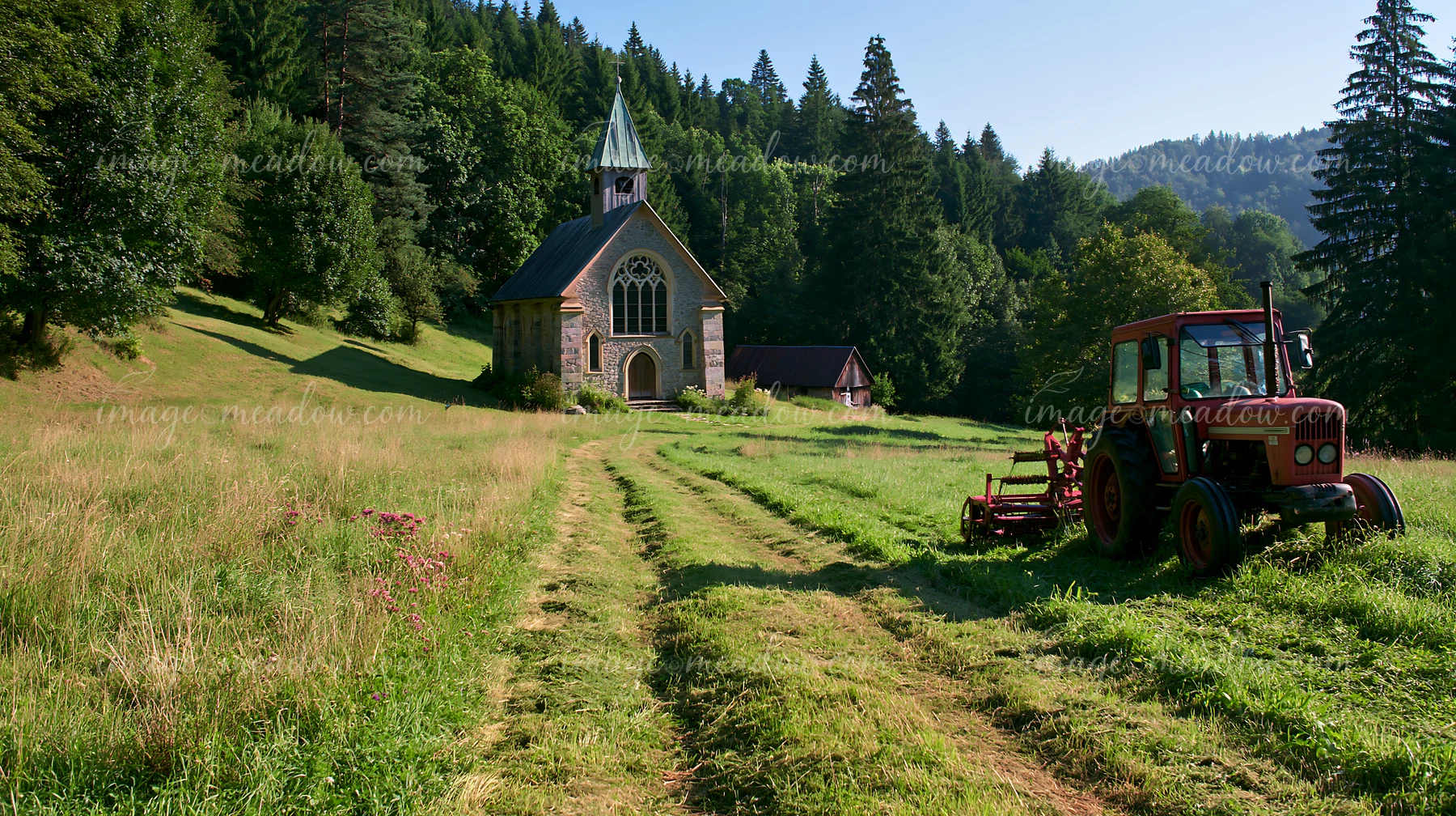 A tractor mows a meadow near a stone church surrounded by trees in the countryside.