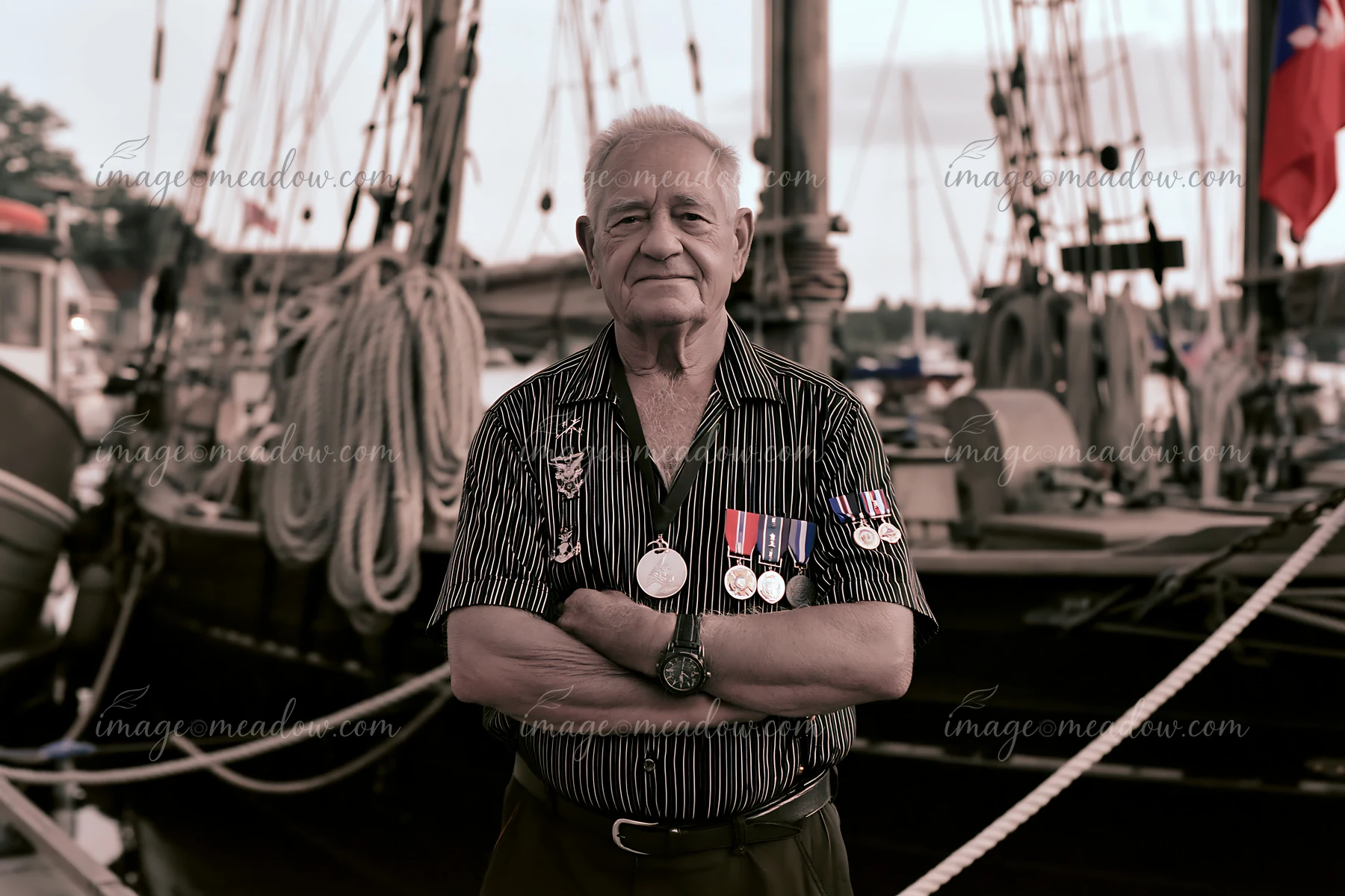 A veteran wearing medals poses confidently at the harbor with ships in the background.