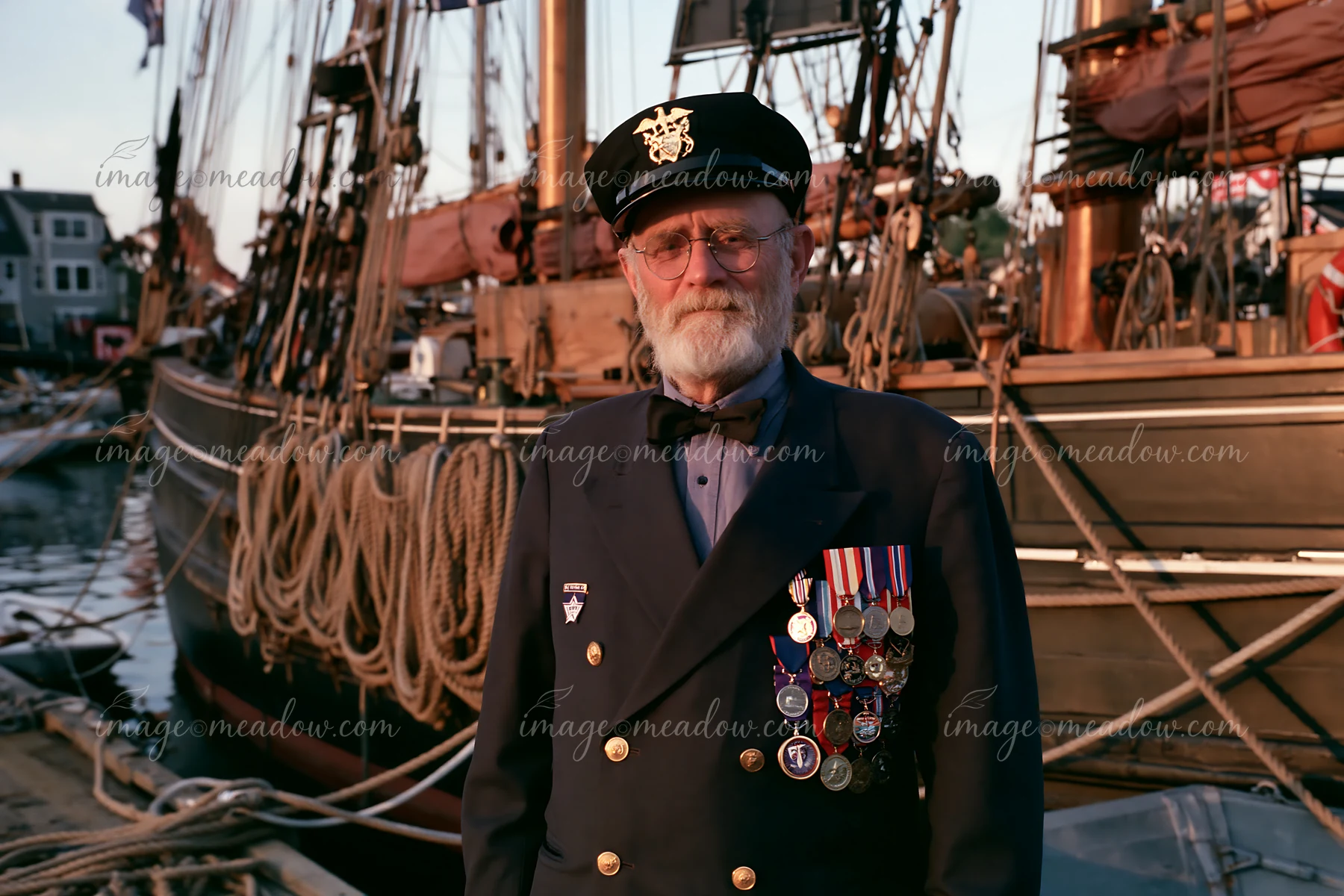 A veteran wearing medals poses near a historic wooden ship in the harbor during golden hour.