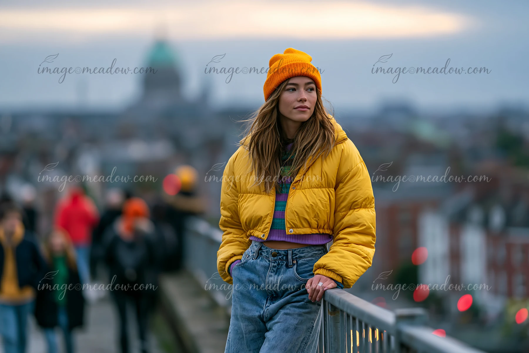 A woman leans against a railing on a rooftop during sunset with people in the background.