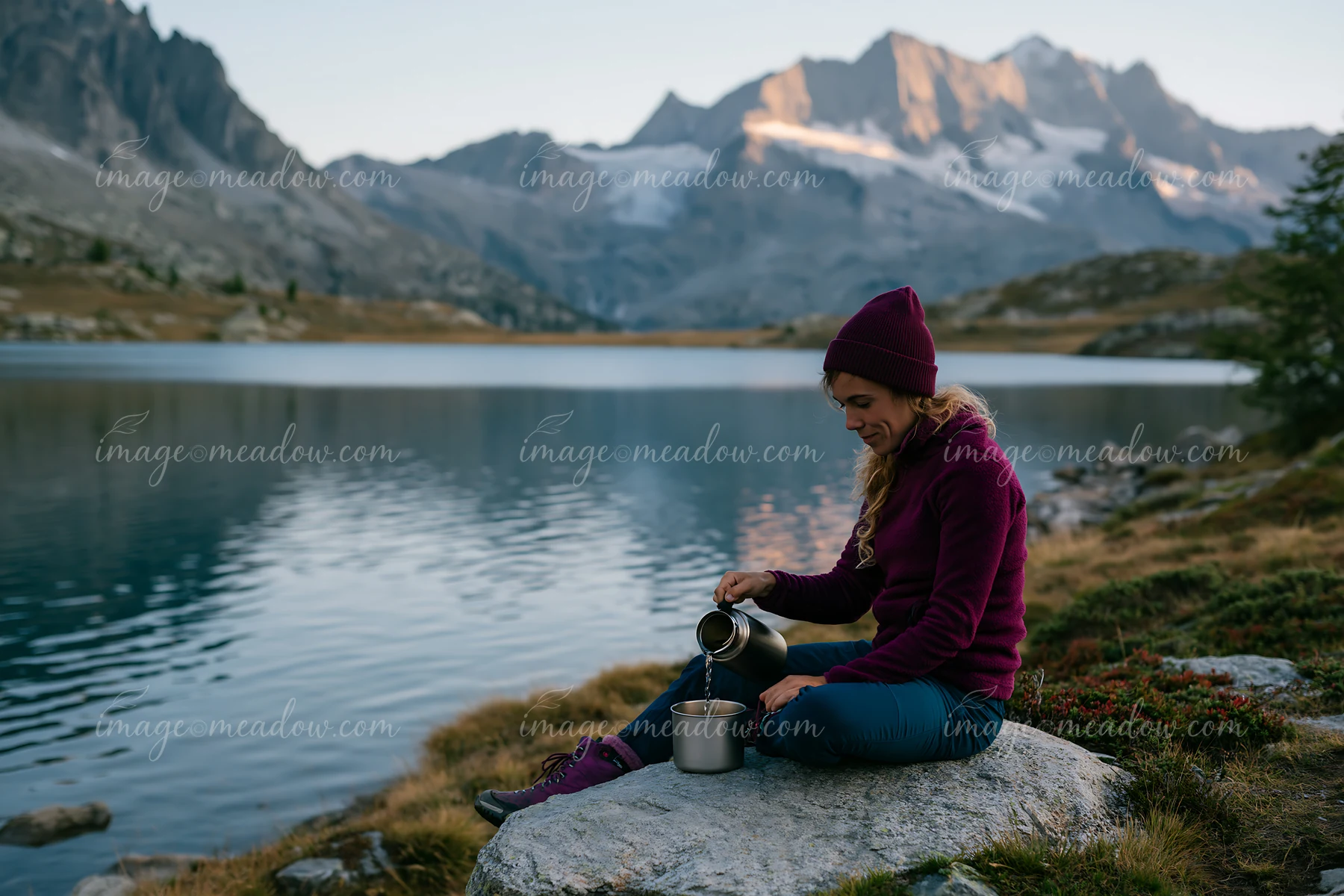 A woman prepares food at a rock by a lake surrounded by mountains.