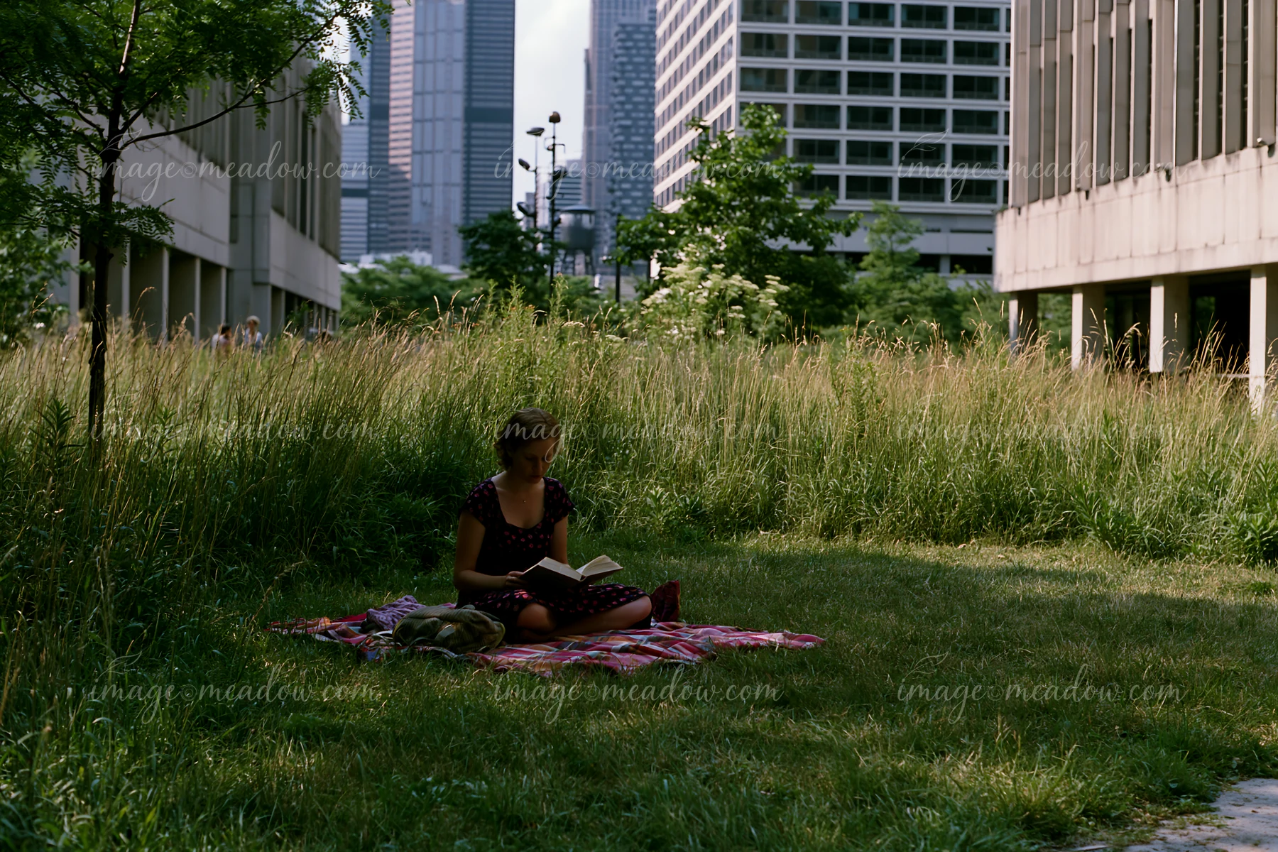 A woman sits on a blanket in a grassy area, reading a book surrounded by tall buildings.
