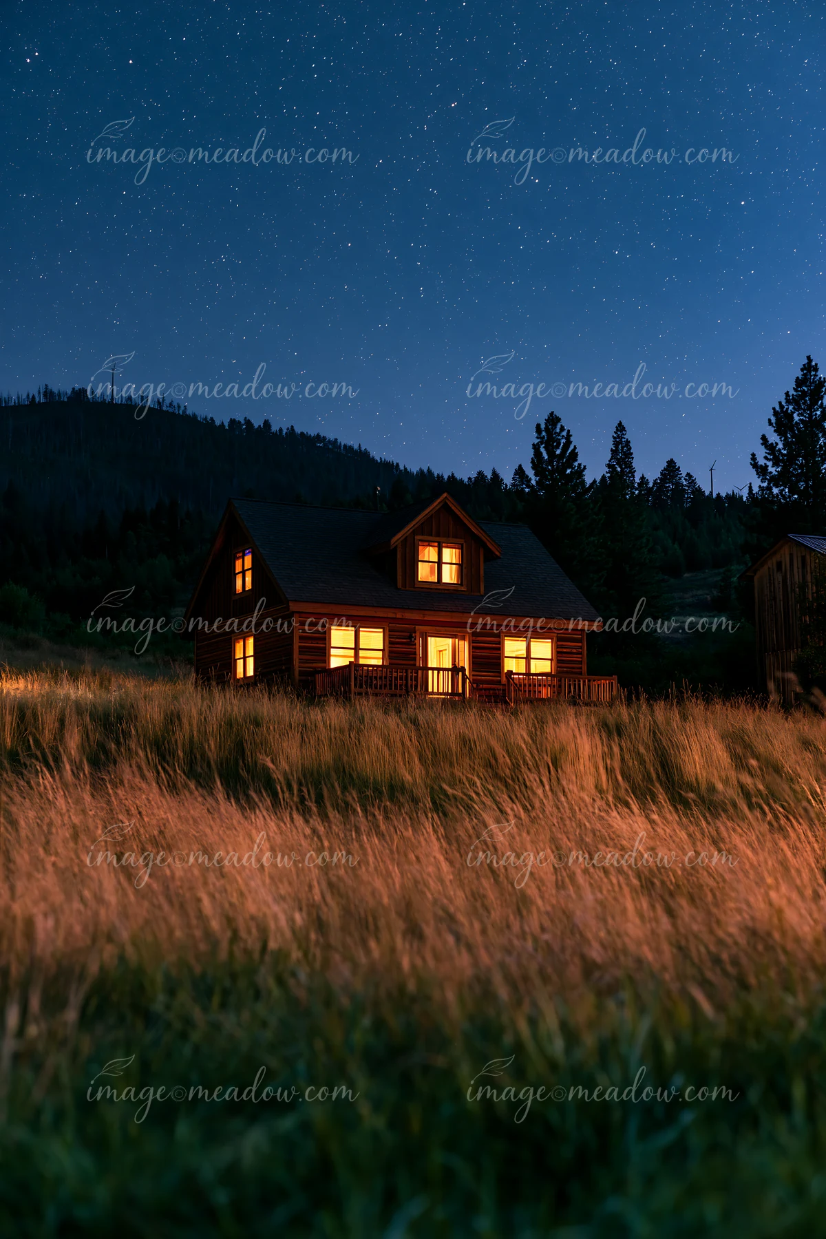 A wooden house sits in a grassy field, illuminated by warm light from its windows.