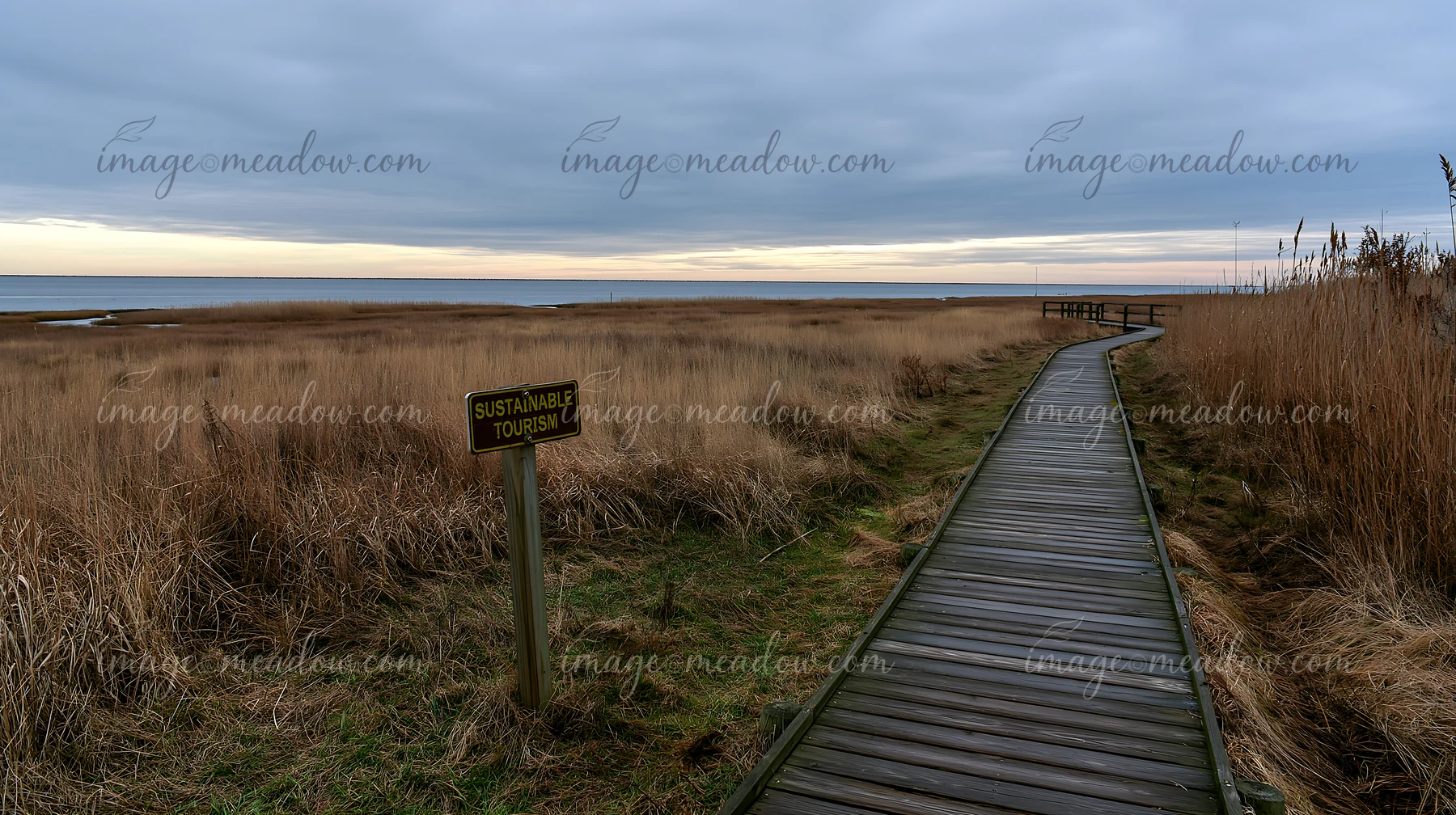 A wooden walkway leads through tall grass to a coastline, where a sign emphasizes sustainable tourism.