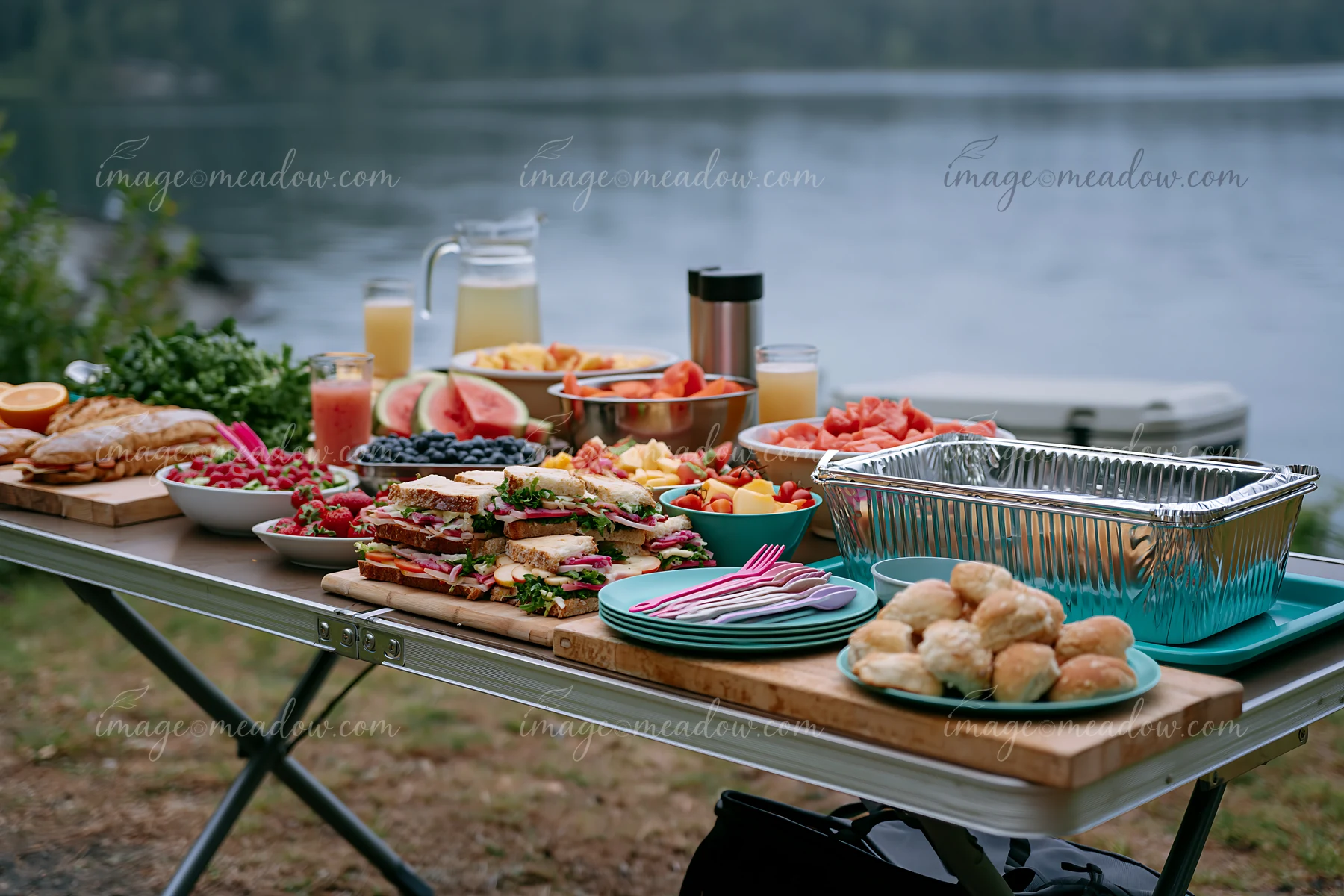 A zero waste buffet table features an array of colorful foods, drinks, and desserts set outdoors near a lake.