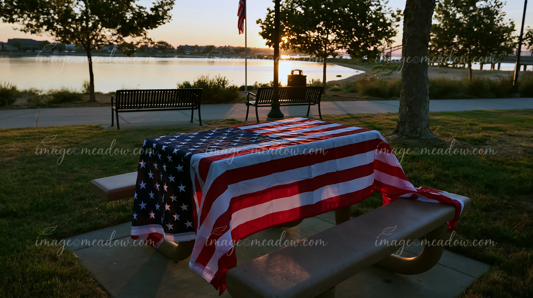 An American flag at dawn covers a table in a park near a calm body of water.