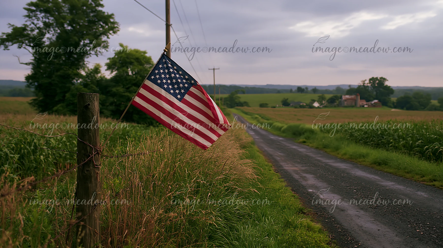 An American flag hangs from a wooden post beside a gravel road in a rural area.