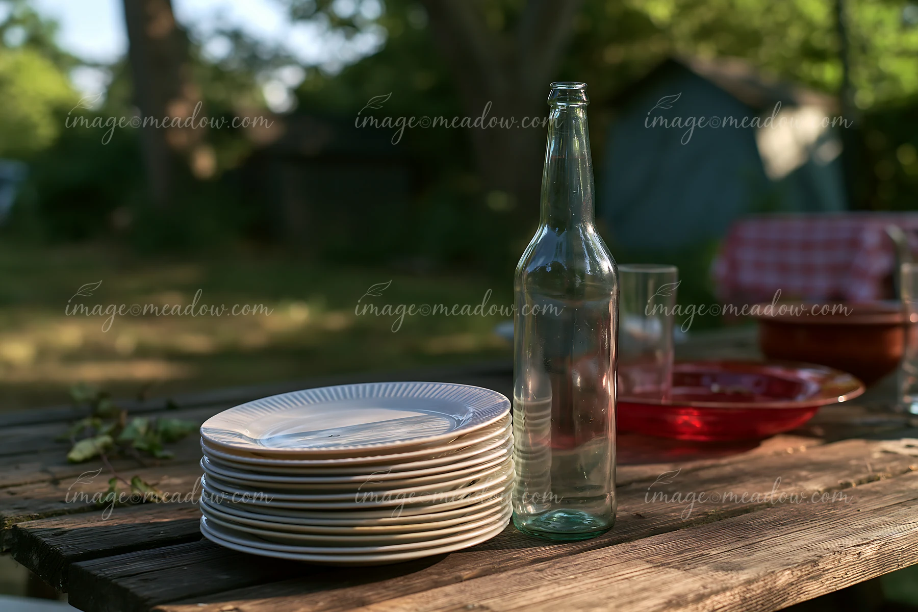 An empty bottle after party stands next to a stack of plates on a wooden table in a garden setting.