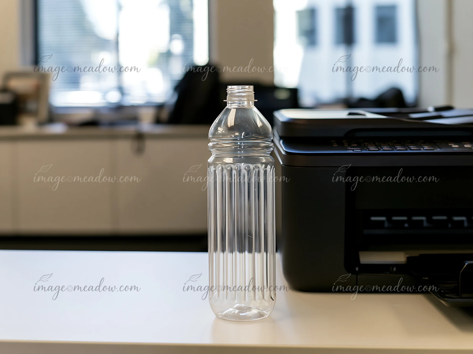 An empty bottle is placed on a desk in an office setting, next to a printer.