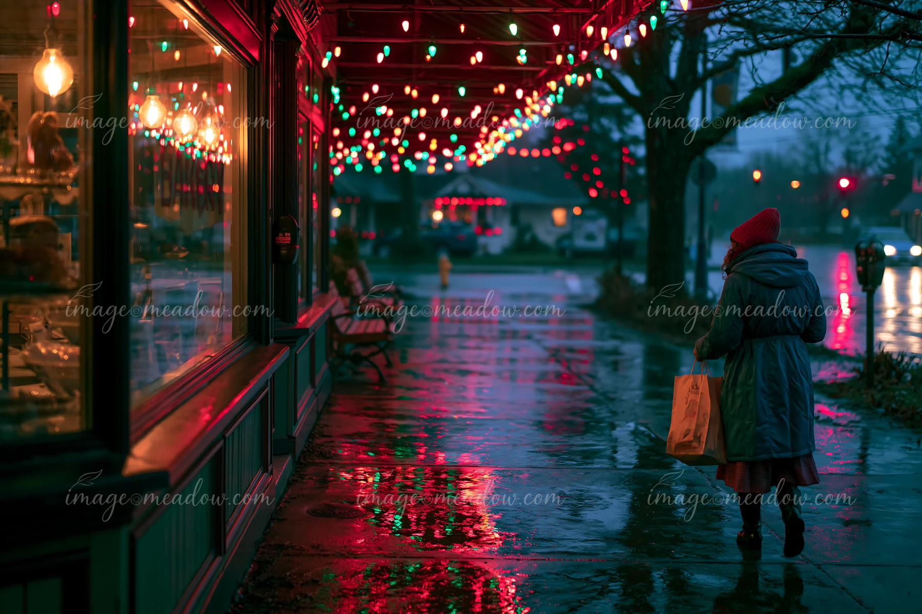 Bundled shopper walks past a decorated storefront as colorful Christmas lights reflect on wet pavement at dusk.