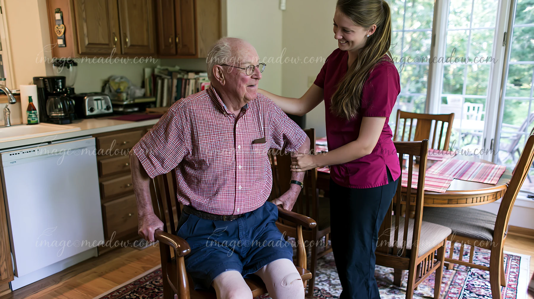 Caregiver supports an elderly man seated in a chair inside a bright home kitchen and dining area.
