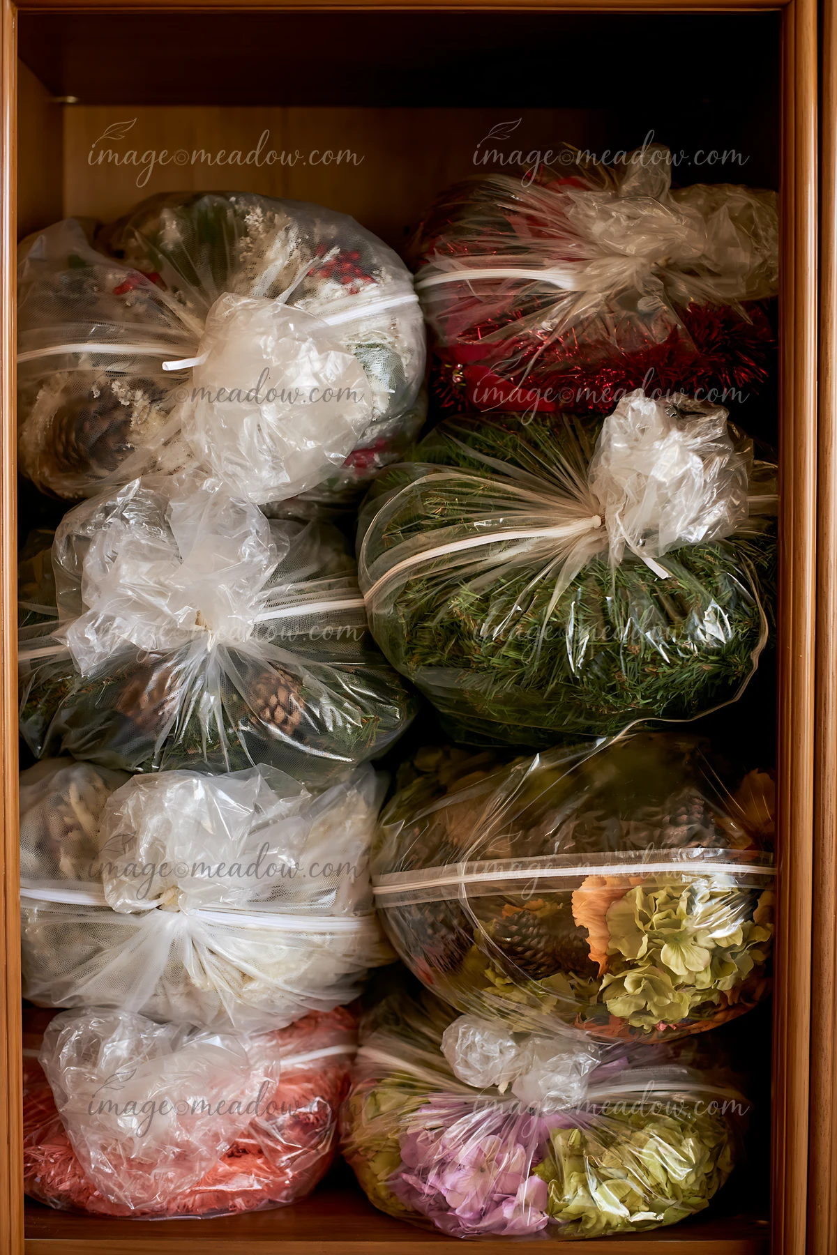Clear bags of Christmas decorations, greenery, tinsel, pinecones, and flowers fill a wooden storage cabinet.
