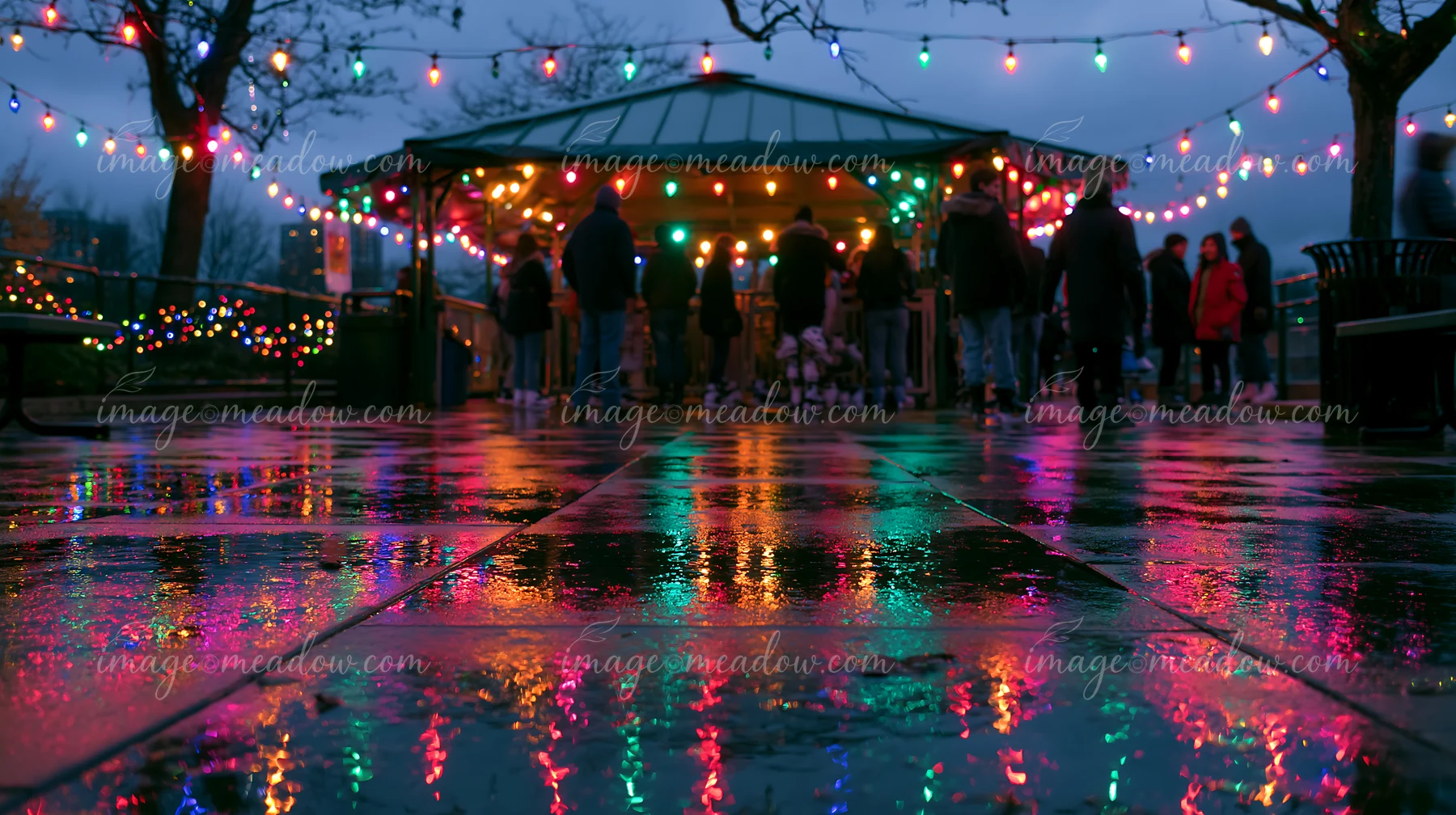 Colorful Christmas lights reflect across wet pavement outside a market pavilion at dusk.
