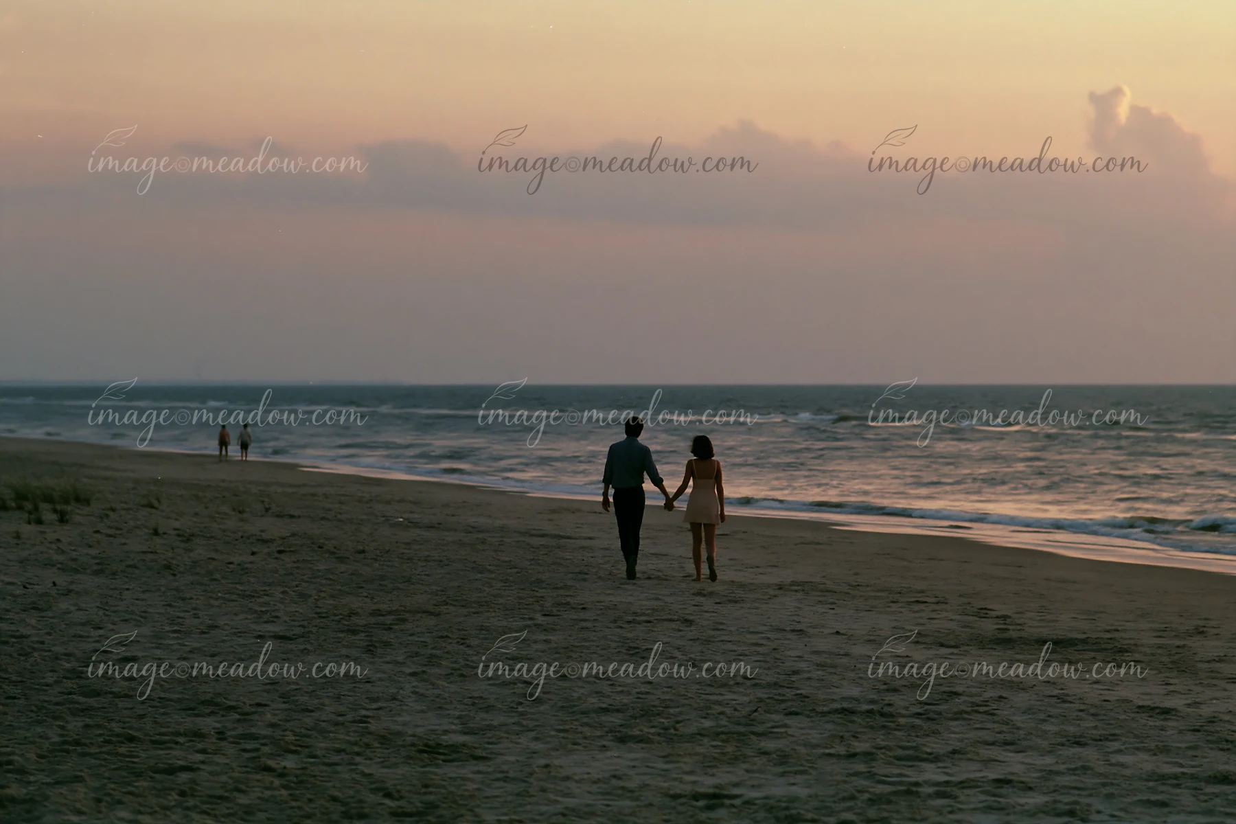 Couple holding hands while walking along a quiet beach beside the ocean at dusk.