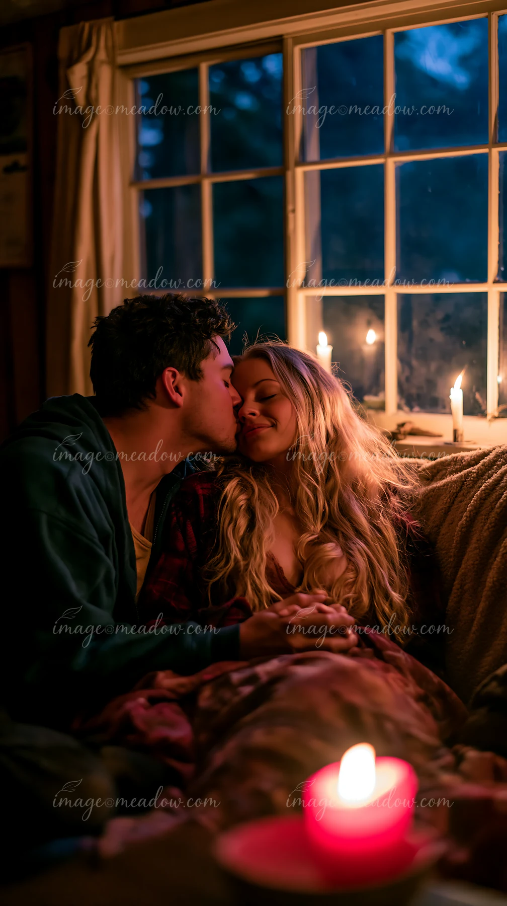 Couple shares an affectionate kiss under a blanket beside candlelight and a window at night.