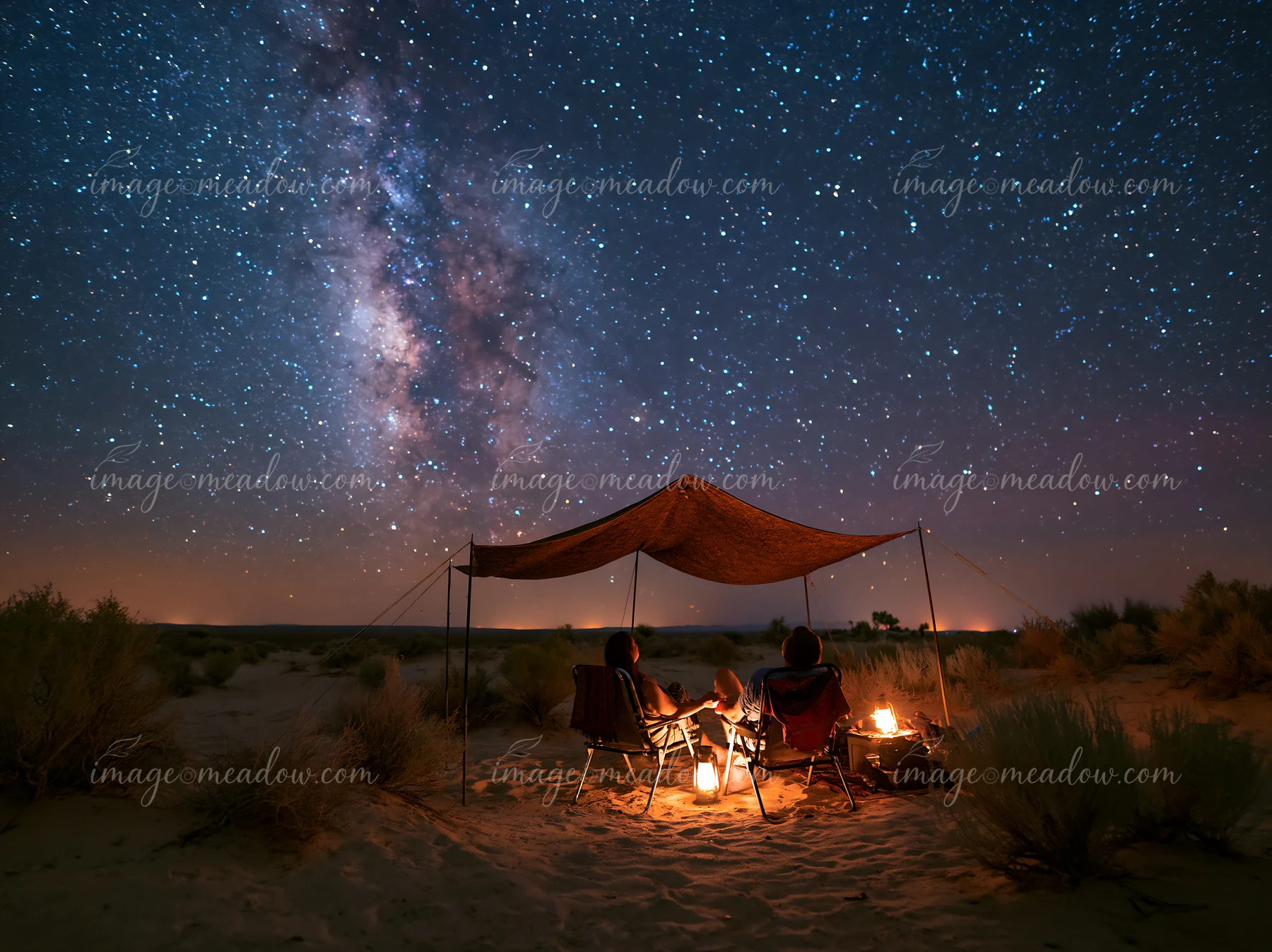 Couple sits beneath a canopy in the desert beside lanterns and a campfire under a star-filled sky.